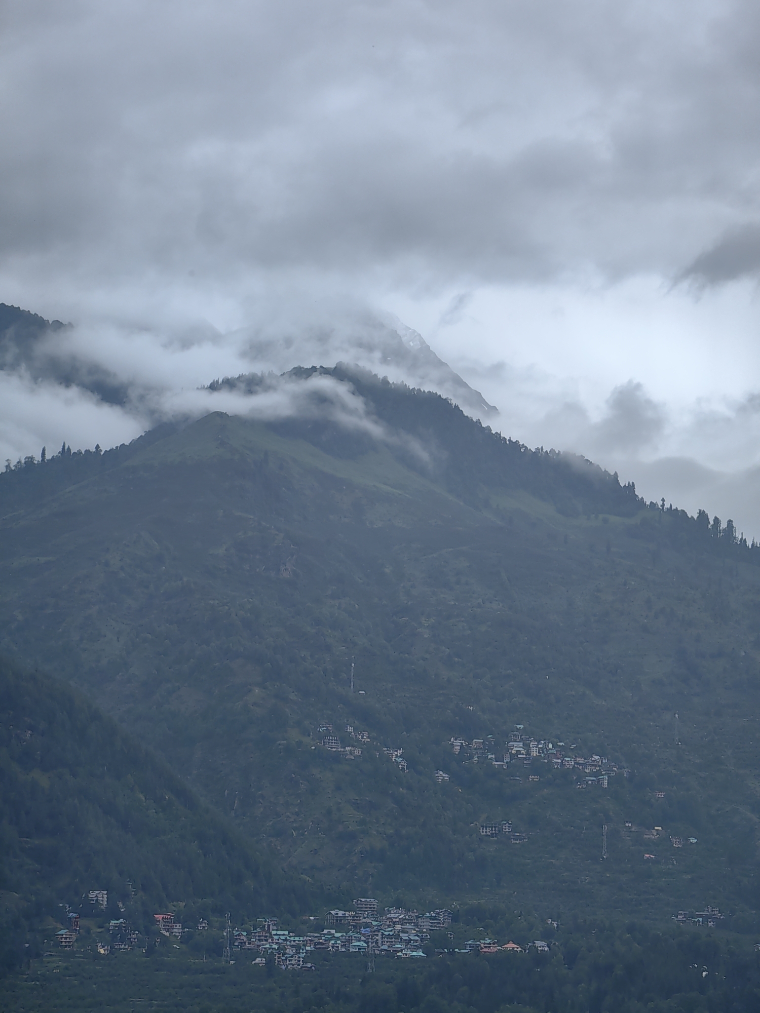 The image depicts a broad view of the mountainous terrain surrounding Vashisht, India. In the foreground, a dense forest covers the lower slopes, transitioning into scattered human settlements built into the hillsides. These villages are characterized by multi-story buildings with predominantly white and light-colored facades, some featuring red roofs, clustered together along winding roads. Telephone poles and small antennas are visible amongst the structures.
The midground is dominated by steep, verdant mountain slopes, heavily forested with pine trees. The texture of the foliage is evident, with the darker green of the trees contrasting with the lighter green of the grassy areas. Some of these slopes appear to be terraced, indicating agricultural activity or land management.
The background features higher mountain peaks, partially obscured by heavy, gray clouds that drape over the landscape. The clouds create a dramatic, moody atmosphere, with some sunlight attempting to break through in diffused shafts. The overall impression is one of a remote, mountainous region, possibly experiencing overcast or rainy weather, with human habitation nestled within the natural grandeur. The image captures a sense of scale and the interplay between the natural environment and human presence in the Vashisht area.