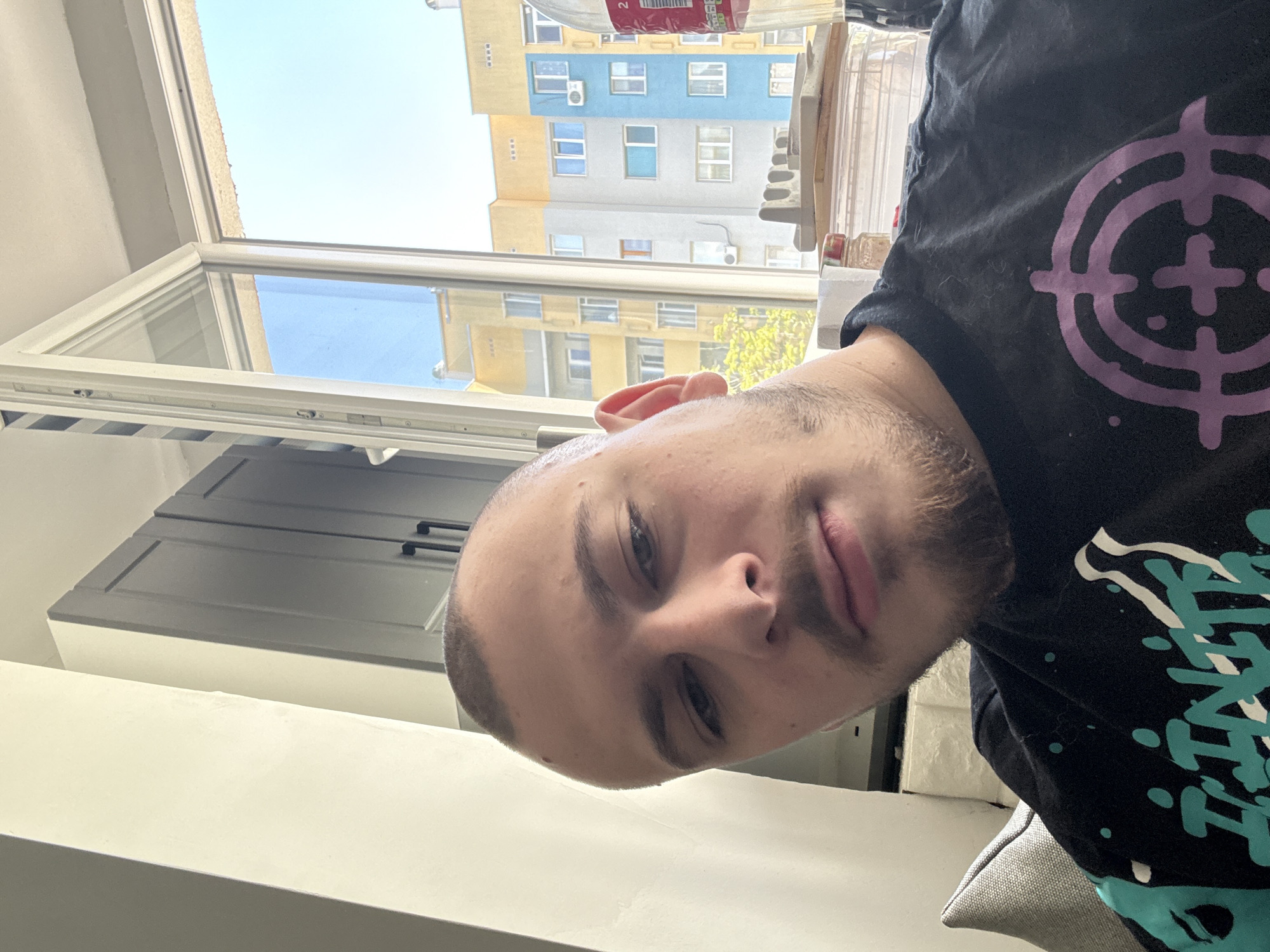 A selfie of a man with a shaved head, wearing a black t-shirt with a purple and white target-like design. The background shows a multi-story building in Stara Zagora, Bulgaria, through an open window. The building is painted in yellow, blue and white. The interior shows grey cabinets, a white wall and other objects.