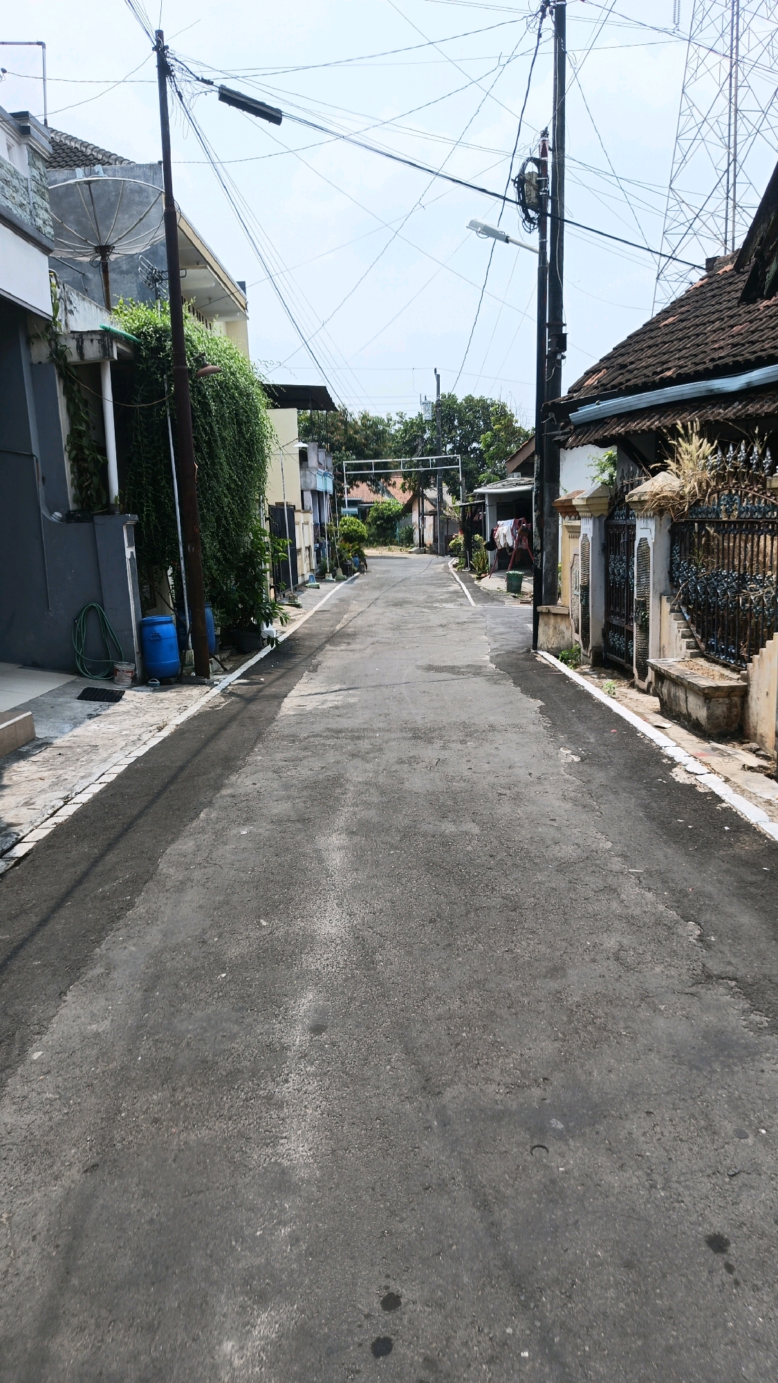 This is a vertical, eye-level, medium shot of a paved residential street in Gedanganak, Indonesia. The street runs from the foreground to the background and is bordered by houses on both sides. The sky is partly cloudy. There are numerous overhead wires and utility poles crisscrossing the scene.
On the left side of the street, a gray and white house with a satellite dish mounted on its side is visible. A dense green vine covers a portion of the house. Near the house, a green garden hose is coiled on the ground next to two blue barrels. A dark pole stands in front of the vine.
On the right side of the street, a house with a dark brown tiled roof and ornate iron gate is visible. A stone bench is situated in front of the gate.
The asphalt street appears worn and shows signs of cracking and some dark stains, likely oil marks. The overall impression is of a simple, somewhat cluttered, and lived-in neighborhood in a tropical climate. No people are present in the image.