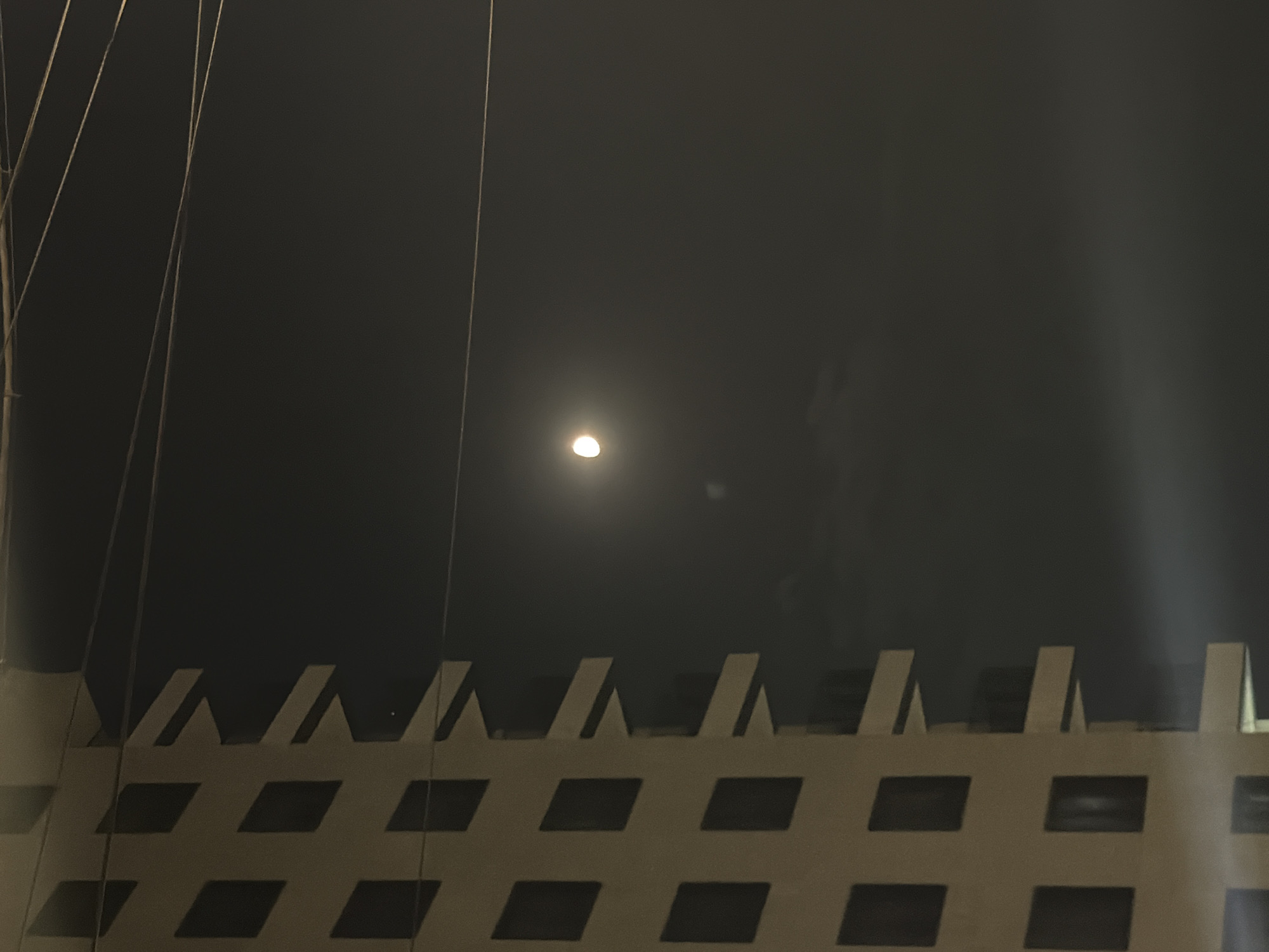 The image captures a nighttime scene in Yerevan, Armenia, featuring the side of a light-colored building with a grid of dark square windows. The building's facade is characterized by repeating rectangular elements that create a textured appearance. To the right of the building, the night sky is visible, dimly illuminated by a bright, possibly full, moon. Several thin electrical wires stretch across the frame, intersecting the sky. A faint, hazy light emanates from the lower right portion of the image, possibly from a streetlamp or other urban light source reflecting off atmospheric conditions or a surface. The overall mood is nocturnal and somewhat abstract due to the lighting and composition.