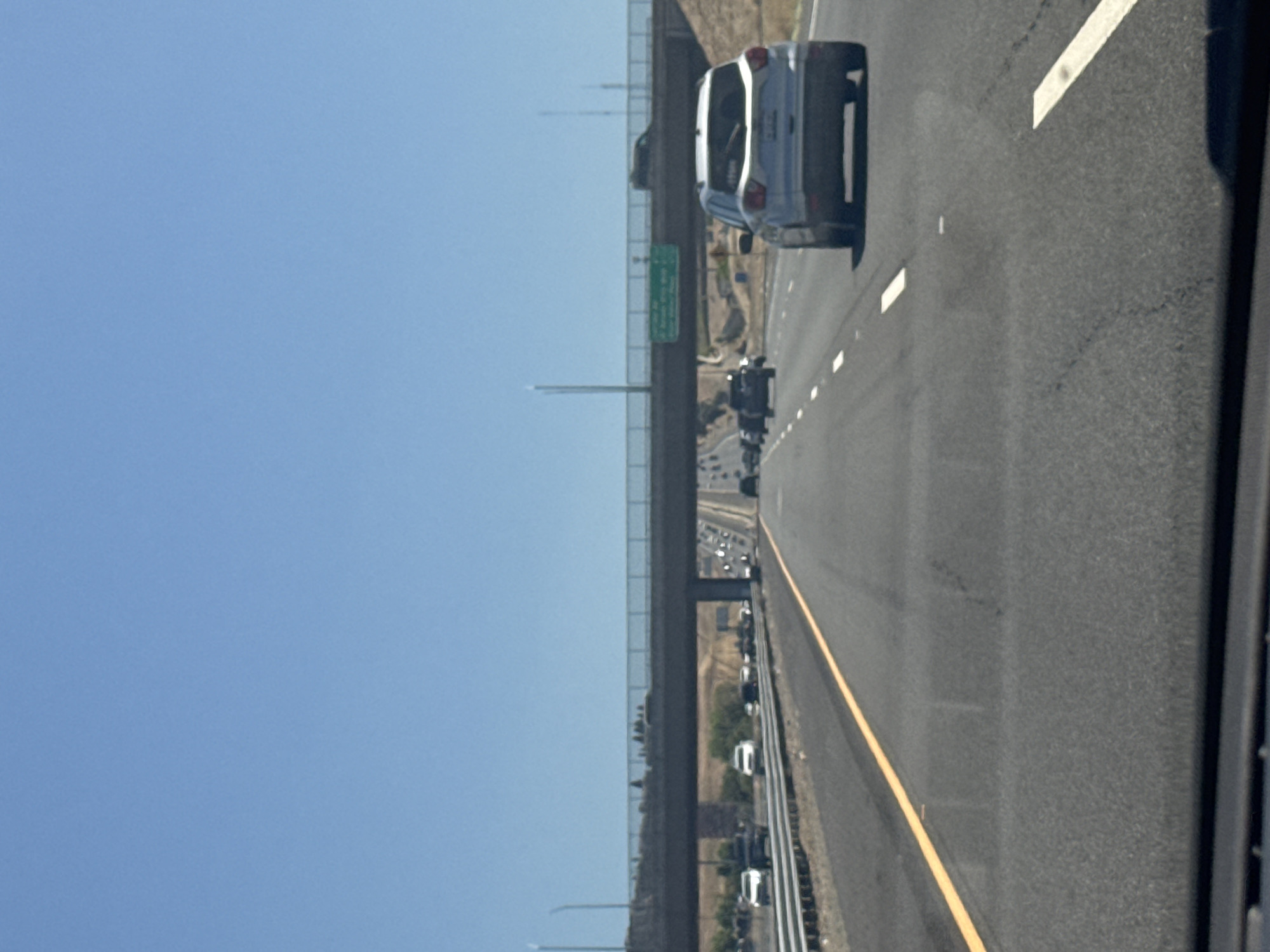 The image shows a view from a vehicle driving on an asphalt highway under a clear blue sky. A concrete overpass stretches across the highway, with traffic visible on both levels. A white SUV is in the foreground, driving in the right lane. Several other vehicles are visible on the highway in the distance. The surroundings appear to be dry and arid, with some vegetation visible on the hillsides. A green highway sign is mounted on the overpass. This scene is consistent with the highway system in Folsom, California, USA.