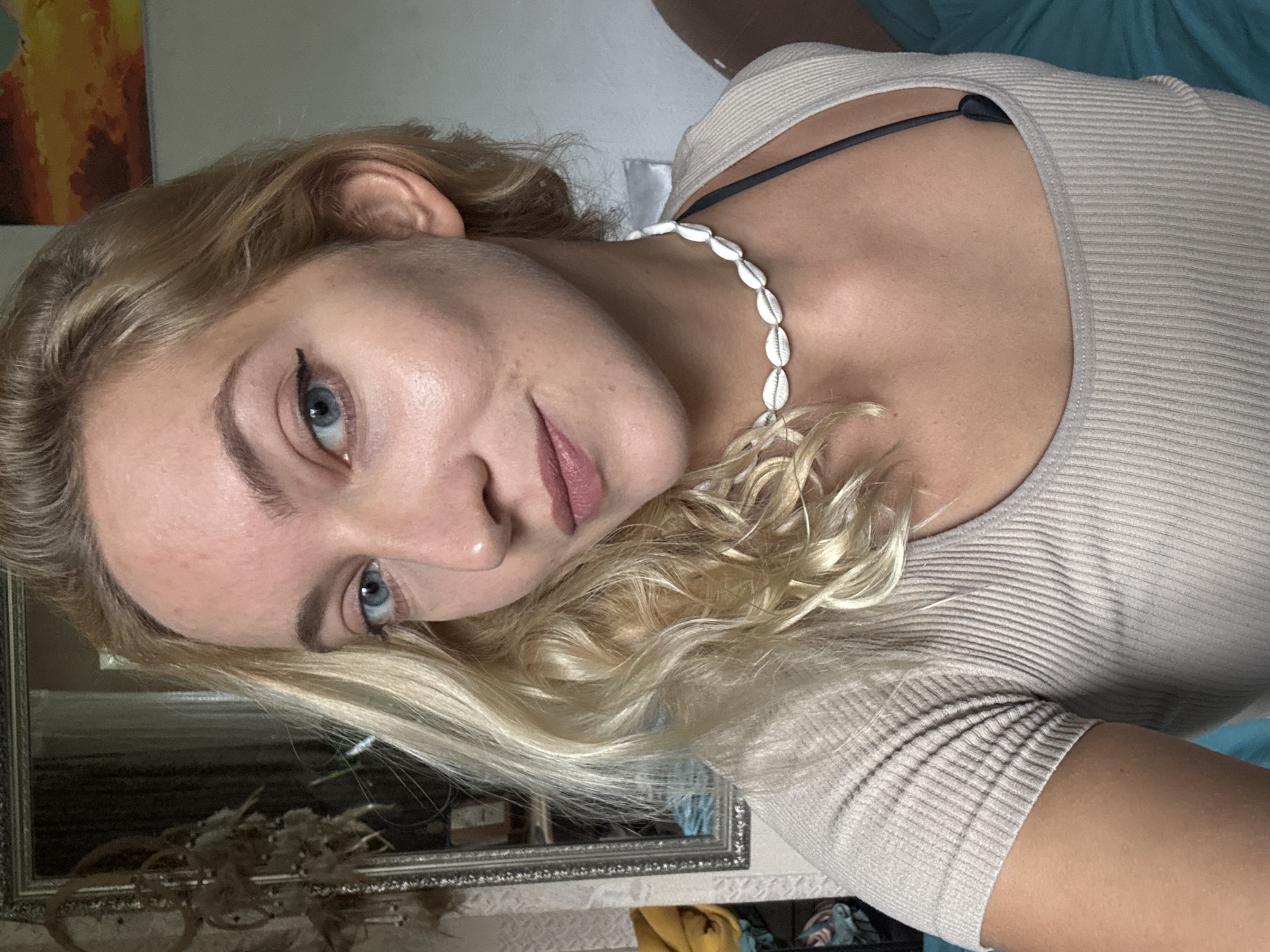 A light-skinned woman with blonde, wavy hair and blue eyes is visible in this image. She is wearing a ribbed, light-colored top and a seashell necklace. The background includes a painting, a mirror with a decorative frame, and what appears to be a portion of a bed or other soft furnishing. The image appears to be taken indoors. The location, Minsk, Belarus, is not discernible from the image.