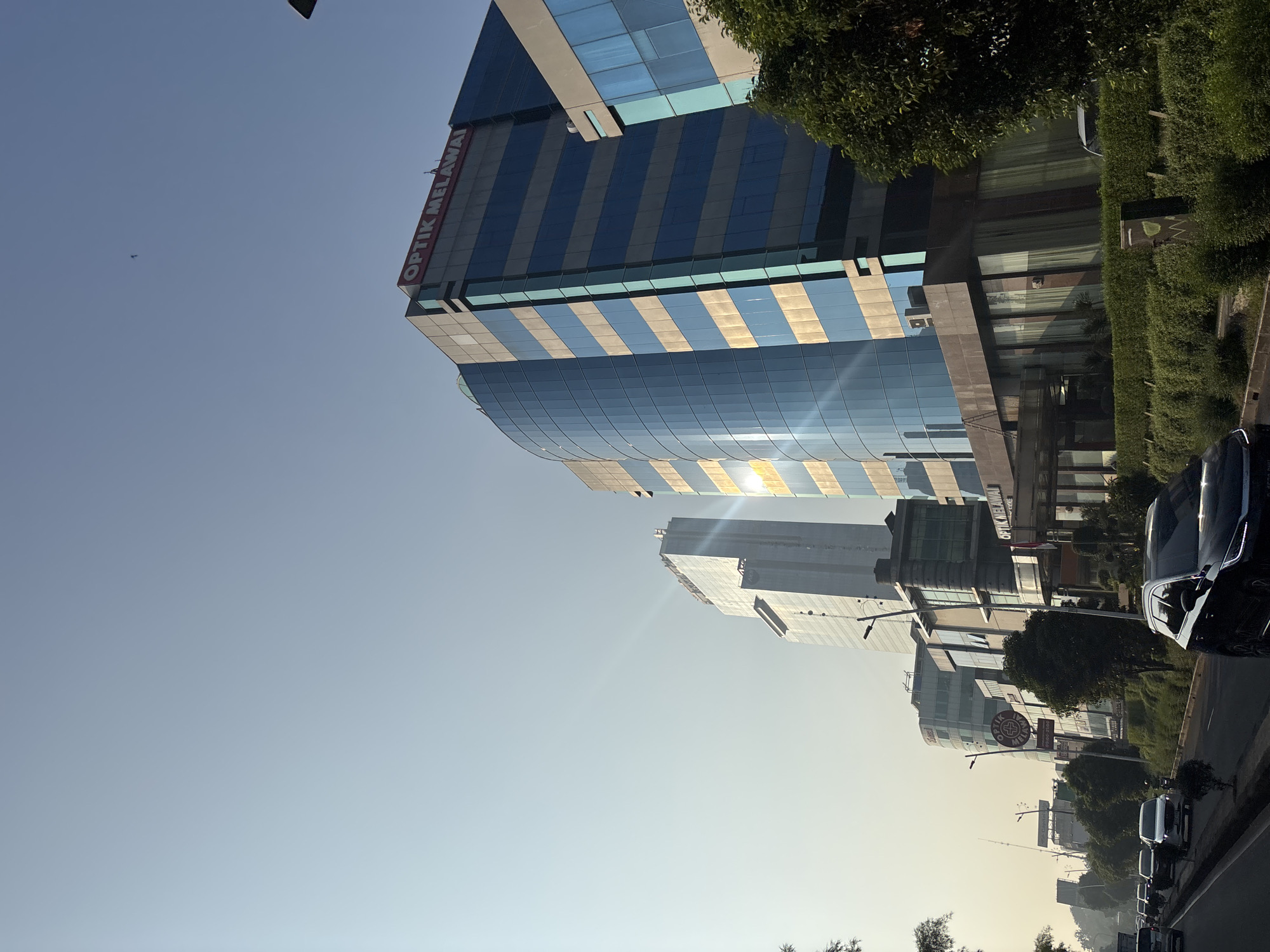This is a low-angle, eye-level, outdoor, eye-level shot of a street in West Jakarta, Indonesia. The image features a street with cars, a building with the words "Optik Melawai" on it, and a clear sky. The sun is shining, and there are lens flares. The building is tall and modern, with blue glass windows and some beige accents. The street is lined with trees and bushes. There are several cars on the road, some of which appear to be in traffic. The overall impression is one of a busy urban environment on a sunny day. The location of this image is on Jl. Daan Mogot, in the Kalideres subdistrict of West Jakarta.