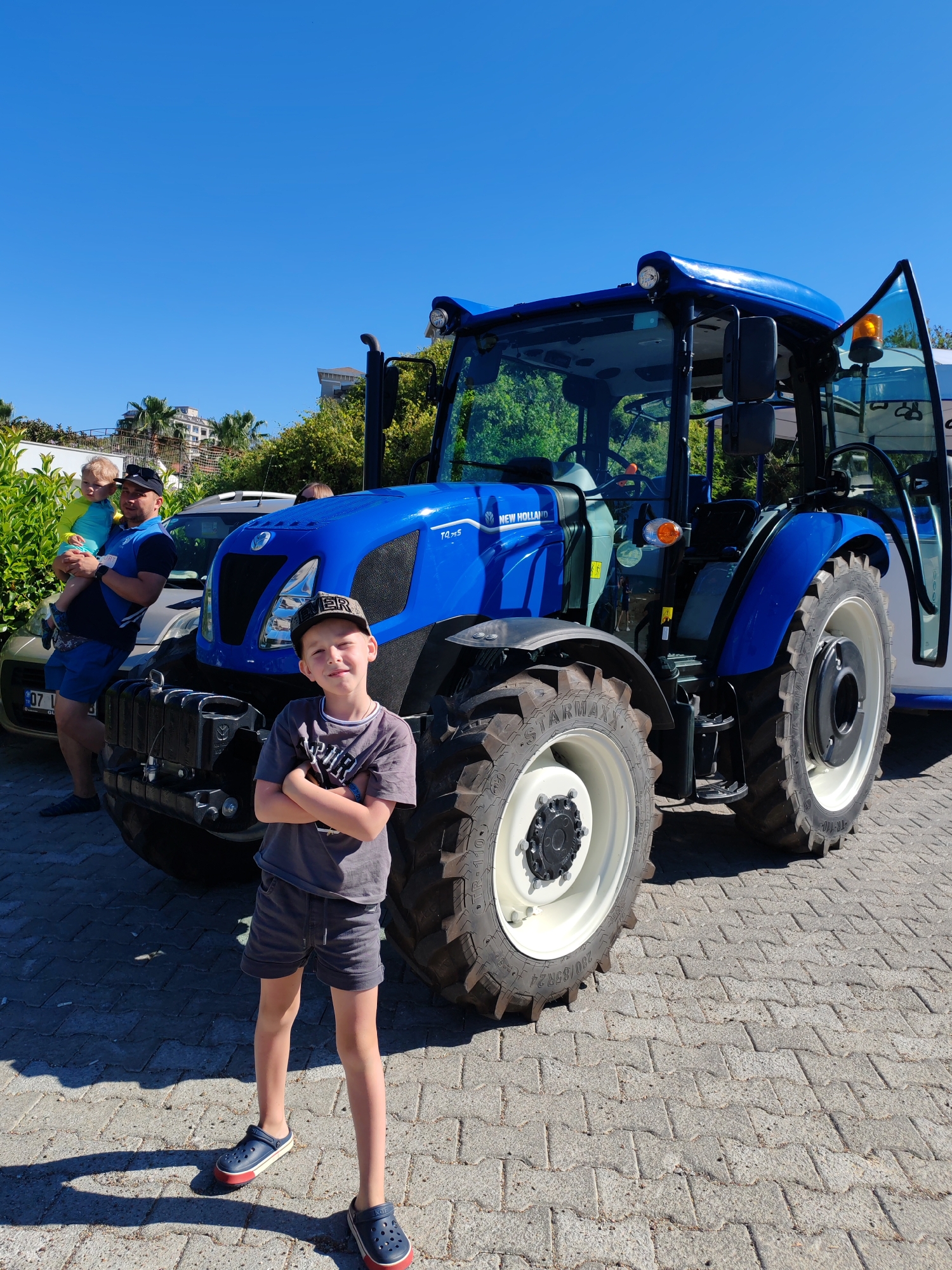 A young boy stands with his arms crossed in front of a blue New Holland tractor. A man holds a young child near a car. The location appears to be in Alanya, Turkey, based on the vehicle license plate. The setting is a paved area near greenery and buildings under a clear blue sky.