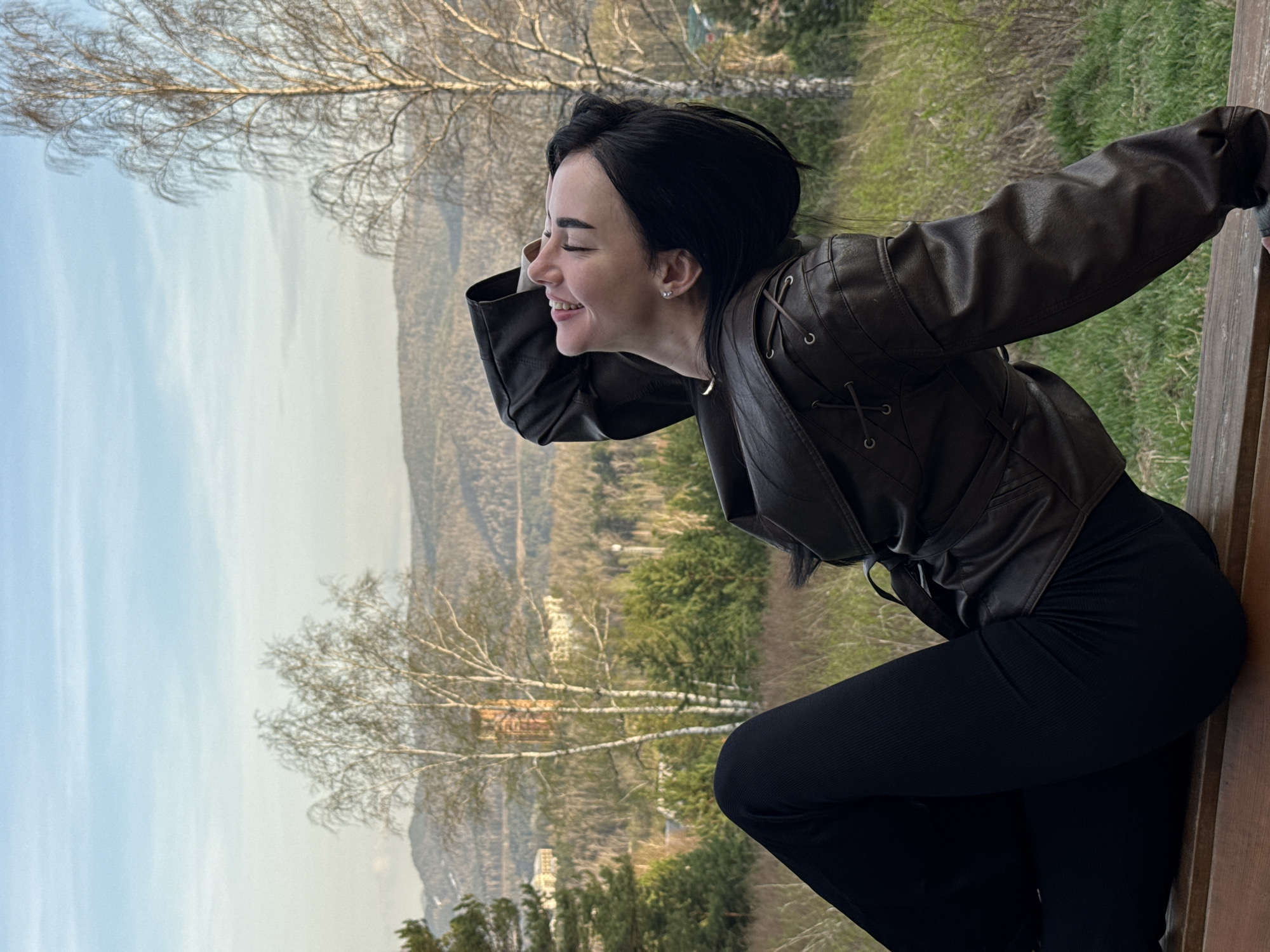 A woman is outdoors, leaning against a wooden railing. She is wearing a dark brown leather jacket and black pants. She is smiling and looking off to the side, towards a landscape of trees and mountains in the distance. The background includes trees with bare branches. The location is Krasnoyarsk, Russia.