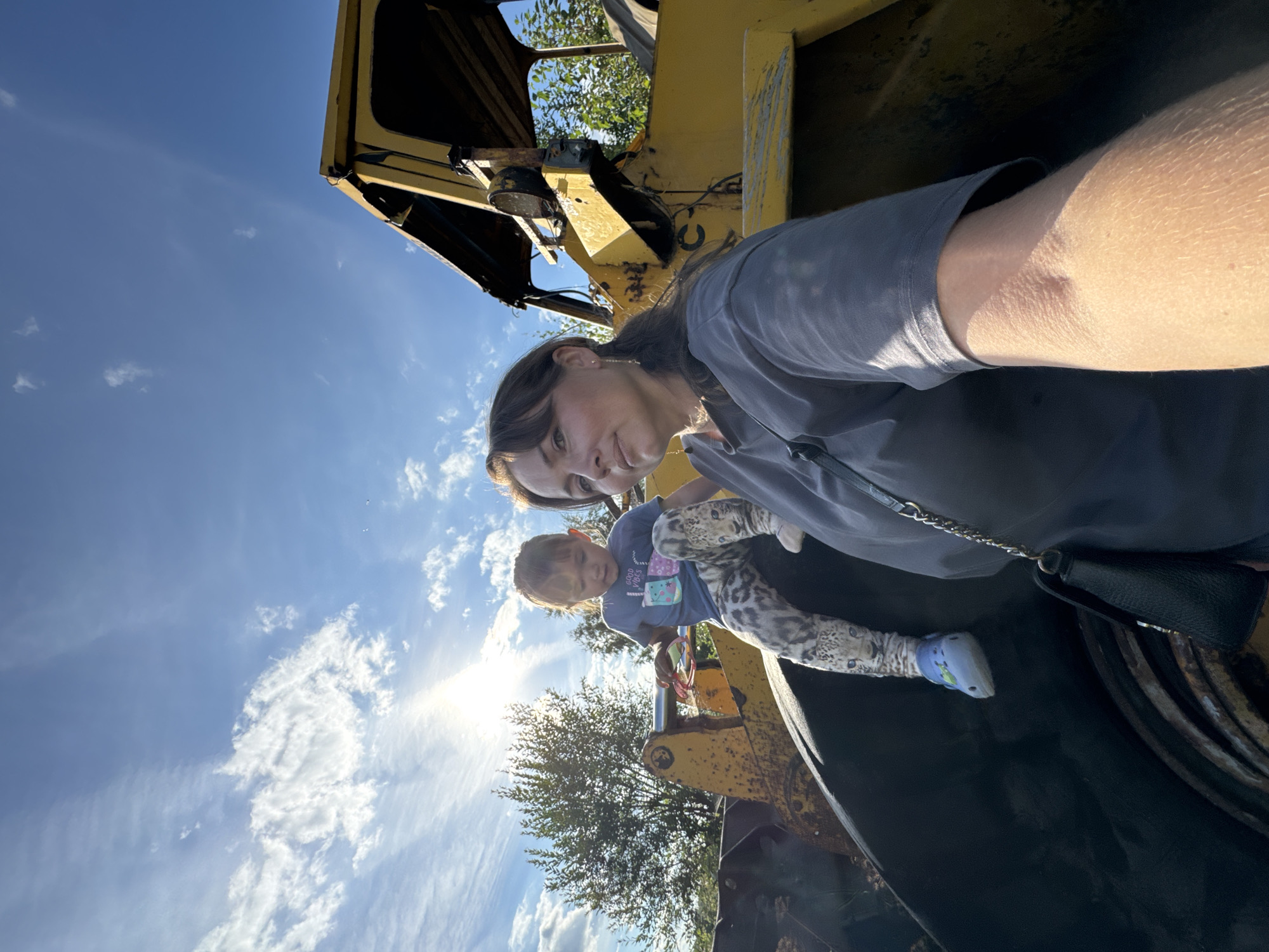 A selfie is taken outdoors in Innolovo, Russia, featuring a woman and a young child. The woman is in the foreground on the right, looking towards the camera. She has dark hair pulled back and is wearing a grey t-shirt and a black purse with a chain strap. Beside her, to the left, is a young child sitting on a large, dark-colored tire. The child is wearing a blue t-shirt with graphics and patterned leggings. Behind them is a large, yellow piece of heavy machinery, possibly a loader or excavator, with a partially visible cab. The sun is shining brightly in the sky, which is blue with scattered white clouds. Trees and foliage are visible in the background. The scene appears to be in an outdoor industrial or junkyard setting.
