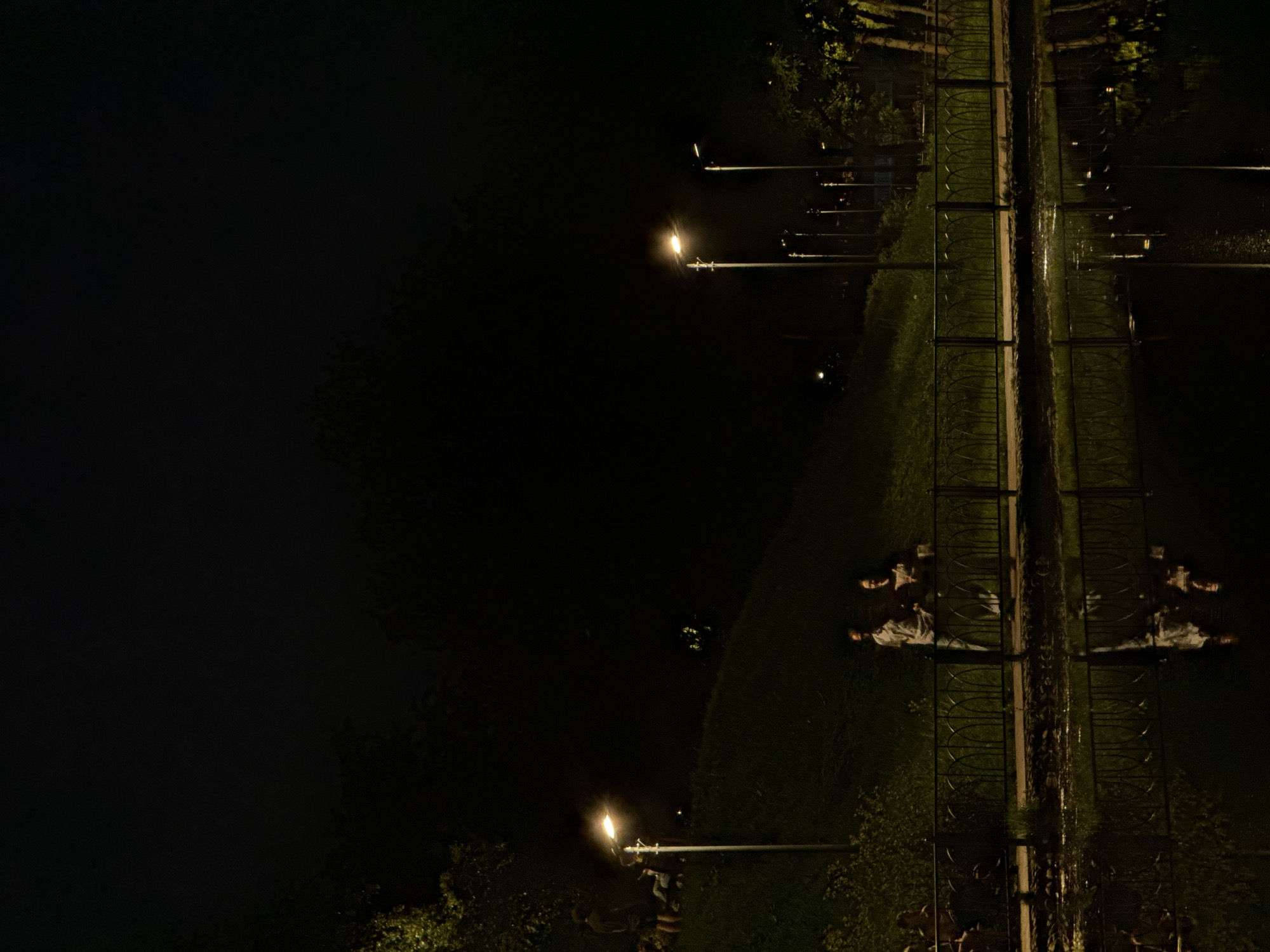 The image depicts a nighttime scene along a path near a body of water, likely in Moscow, Russia. The right side of the image shows a lighted path with a fence running along it. There are lampposts illuminating the path and grassy areas on either side. Reflections of the path, fence, and lampposts are visible in the water, creating a mirrored effect. The left side of the image is dark and shows the body of water, which may be a river, pond, or canal. Two figures are laying on the grass, perpendicular to each other. One figure is wearing light clothing and a hat or mask.