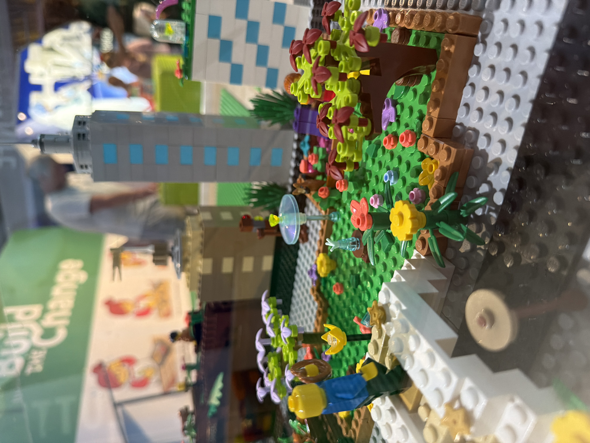 The image shows a detailed LEGO model of a cityscape, specifically a section that appears to be a park or garden area adjacent to a high-rise building. The model features miniature LEGO figures, trees, flowers, and buildings constructed from LEGO bricks. In the background, a large green banner with white lettering that reads "Build the Change" is visible. There are also blurred figures of people in the background, suggesting this is a public display or exhibition, possibly at a museum or event. The overall scene depicts a vibrant and imaginative urban environment constructed from toy bricks. Without further information, it is impossible to determine the specific location within Fort Lauderdale, Florida.