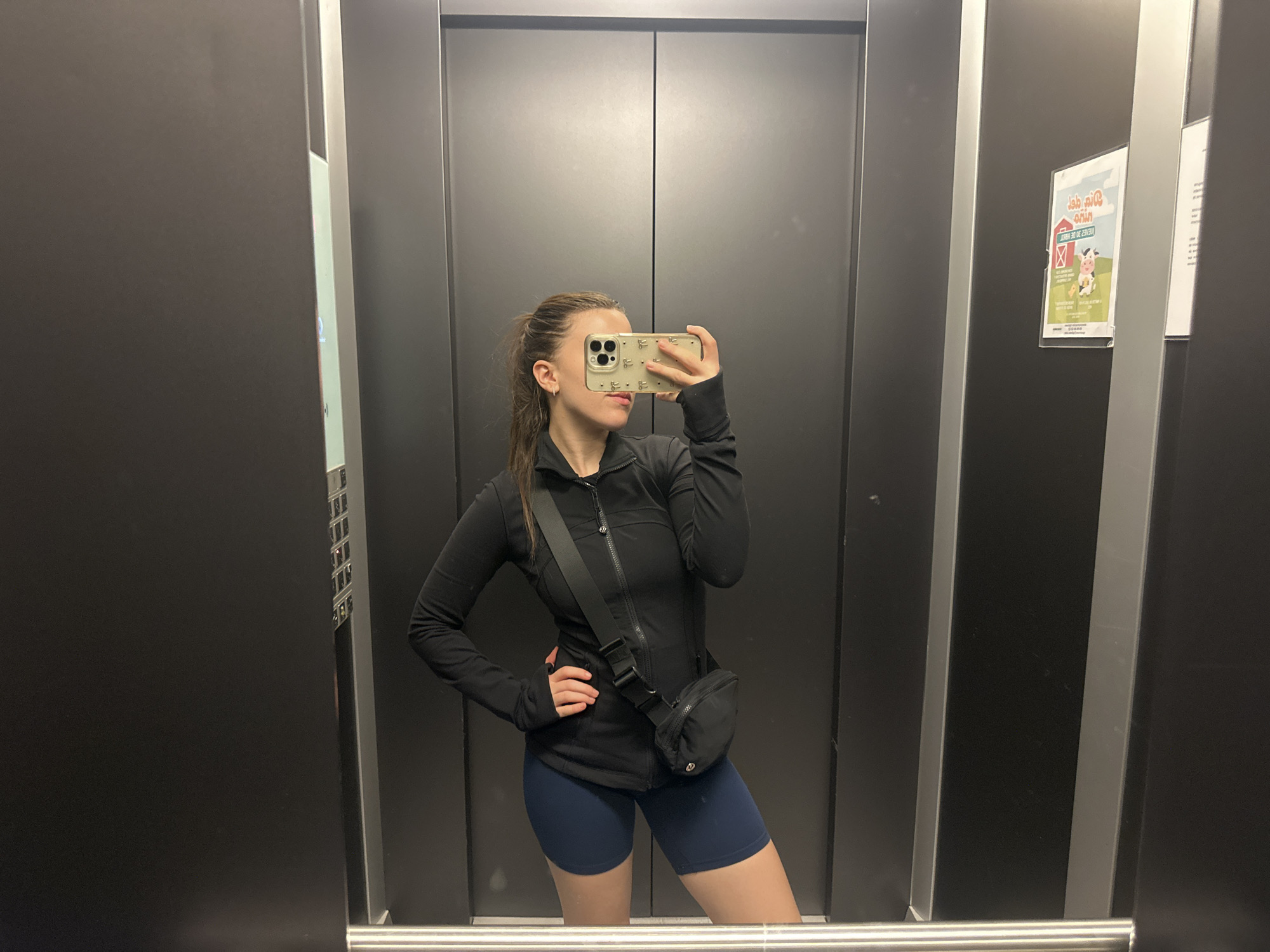 A young woman is captured in a mirror selfie inside what appears to be an elevator. She stands in the center of the frame, holding a smartphone with a light-colored, patterned case up to her face, obscuring most of her expression except for her chin and mouth. Her hair is tied back in a ponytail. She is dressed in athletic wear, consisting of a black long-sleeved, zip-up top and navy blue cycling shorts. A black fanny pack or sling bag is worn across her body, resting on her hip.
The setting is an elevator with dark gray, reflective walls and prominent silver vertical strips, creating a sleek appearance. A silver horizontal railing is visible at the bottom of the frame, reflecting the floor. To the right, on the elevator wall, a colorful poster or sign is affixed. While much of the text is mirrored and partially obscured, the words "ARDI 30 DE JULIO" are discernible, likely indicating a date. The poster features illustrations of a red barn, a white cow with black spots, and a yellow chick, suggesting a farm or animal-related theme. The image provides no specific time of day or weather cues, and the woman's expression is neutral, focused on taking the photo. The provided context indicates the location is Huixquilucan de Degollado, Mexico.