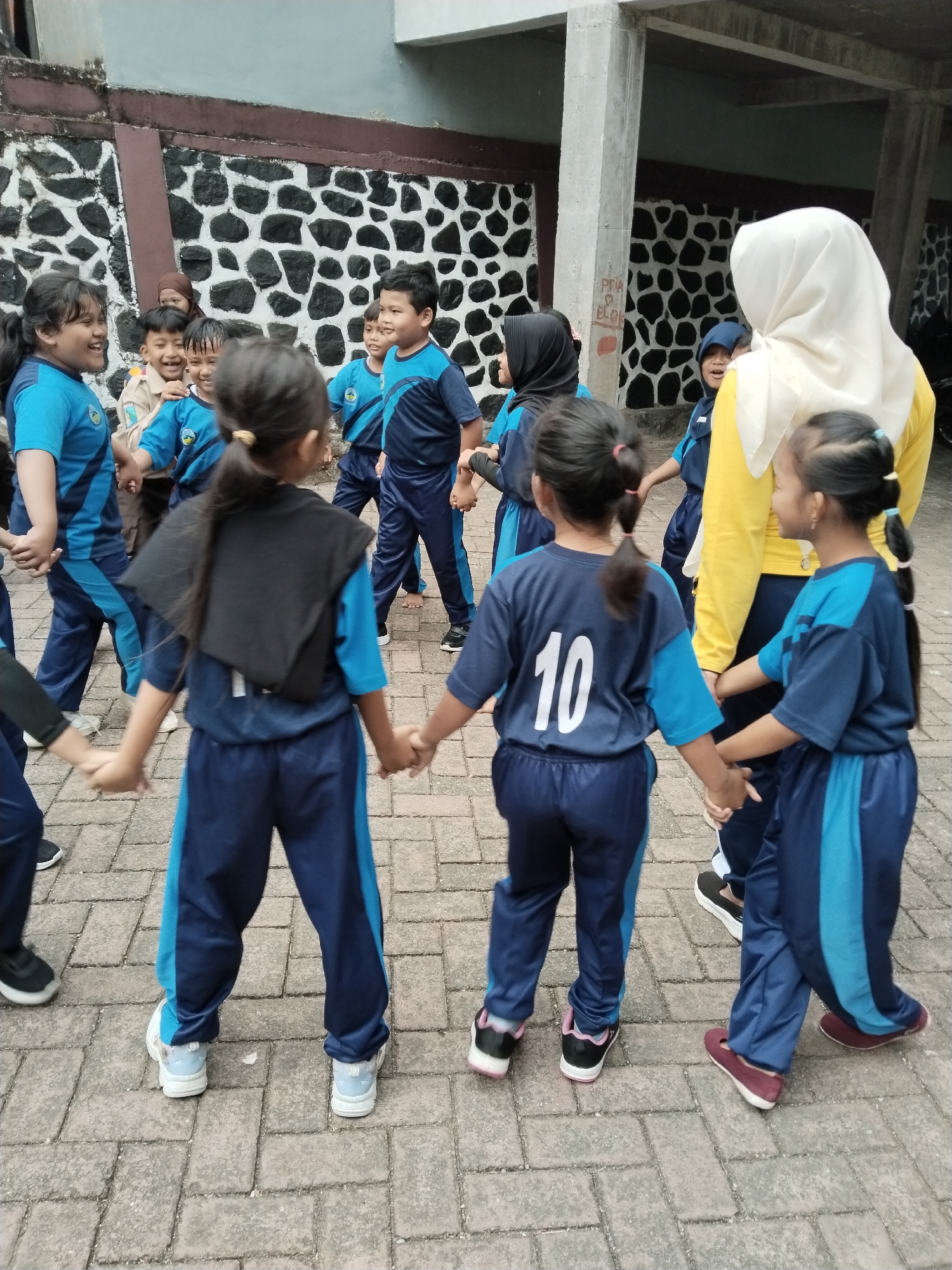 In Bendoroto, Indonesia, a group of children, likely students, are engaged in a circle dance outdoors. They are all wearing matching blue and dark blue athletic uniforms with lighter blue accents. A woman wearing a yellow long-sleeved shirt and a white hijab stands to the right, observing the children. The children are holding hands, forming a circle. One child in the foreground has the number "10" printed on the back of their shirt. The ground is paved with light gray interlocking bricks. Behind the children, a wall with a black and white stone pattern is visible, and to the right, a concrete pillar suggests the presence of a building or structure. The overall scene depicts an outdoor activity involving children and an adult, likely in a school or community setting.