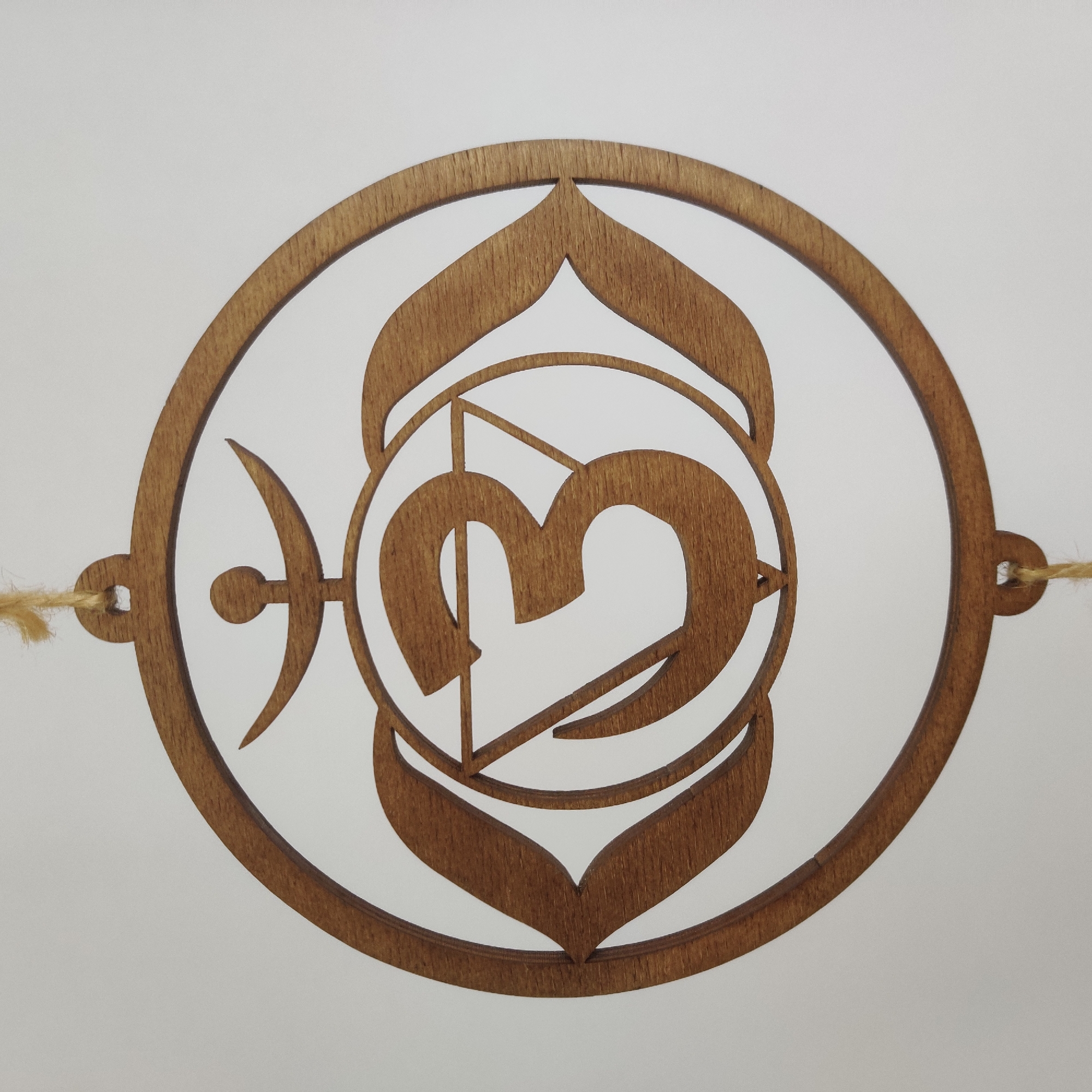A wooden circular ornament hangs against a plain white background. The ornament is intricately carved with the symbol for Om, also known as Aum, a sacred sound and spiritual icon in Indian religions. Within the outer circle, a stylized representation of the Om symbol is present, featuring a prominent large "3" like curve, a crescent moon with a dot above it, and a triangular shape. A loop at the top and bottom of the outer circle suggests it is designed to be hung, with visible strands of twine attached to these loops. The wood appears to be a light to medium brown color with visible grain. There is no indication of its location in Yekaterinburg, Russia.