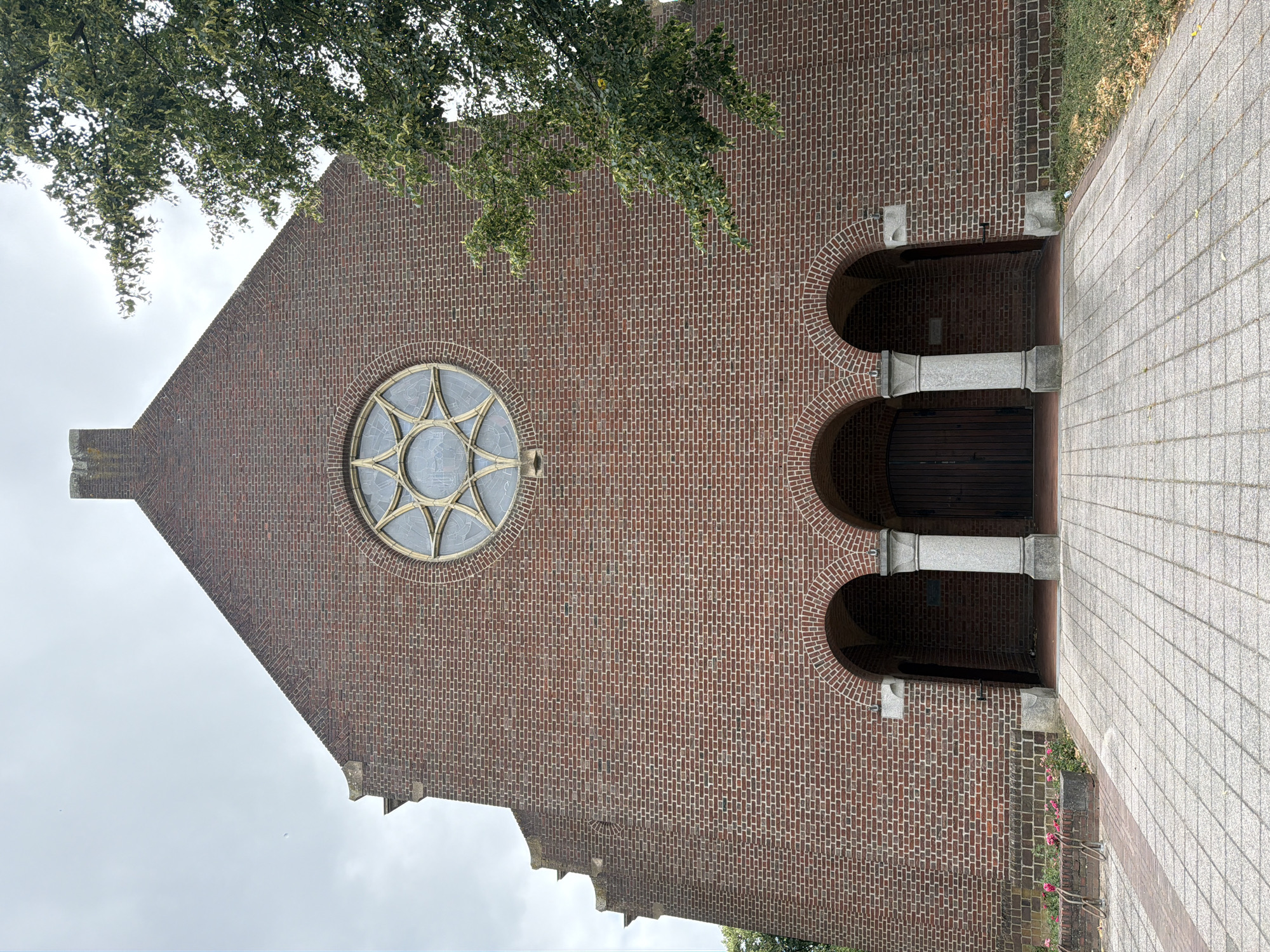 The image shows the facade of a brick church in Melick, Netherlands. The church has a large rose window with a star pattern above a set of three arched entrances. The entrances are supported by stone columns. A tree branch with green leaves extends into the frame from the right side of the image. The ground in front of the church is paved with grey square tiles. In the lower left corner, there are some pink flowers and a bicycle rack. The sky is overcast.