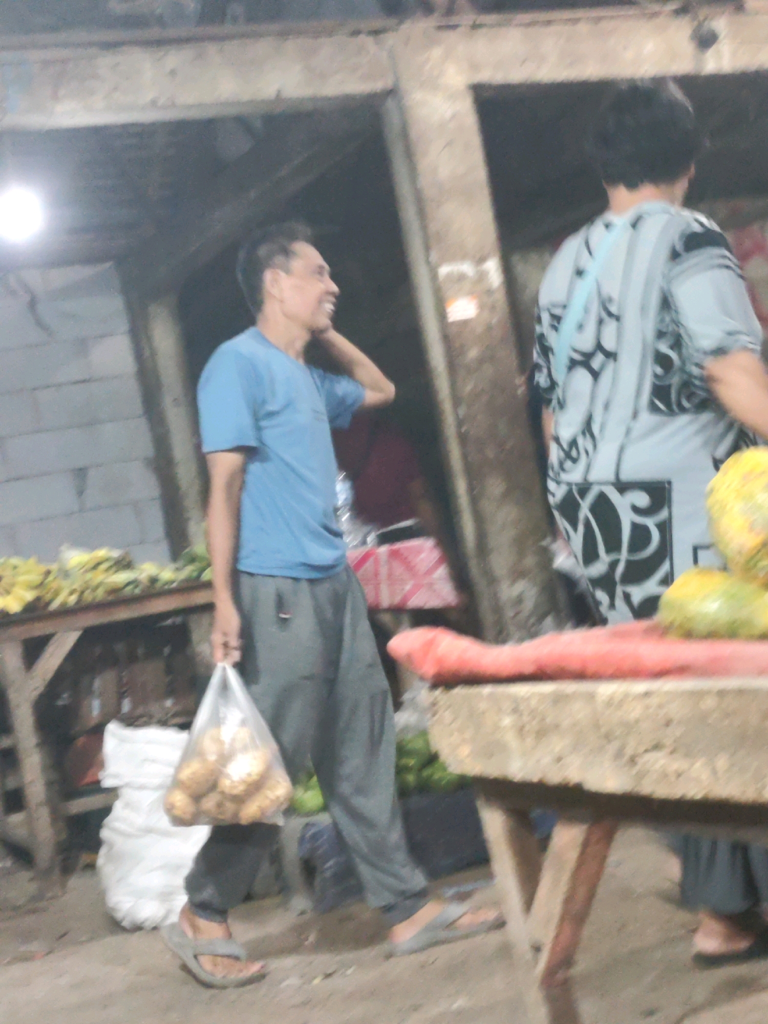 The image depicts a market scene. A man in a blue t-shirt and grey pants is walking, holding a plastic bag filled with what appear to be potatoes. He is wearing flip-flops. To his right, another person is partially visible, wearing a patterned shirt. In the background, there are stalls with produce, including bananas on a wooden table, and various sacks. The overall environment suggests an outdoor or semi-covered market. The lighting is dim, with a bright light source visible on the left. The market appears to be located in Karawang, Indonesia.