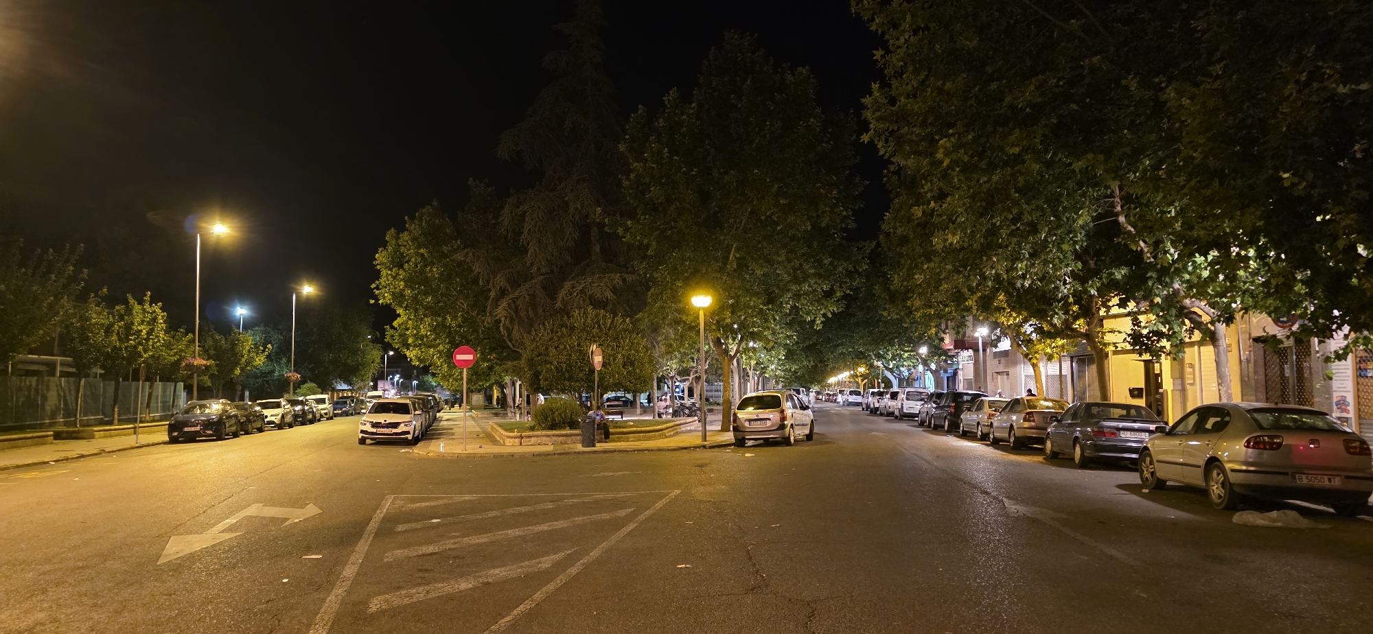 The image depicts a street scene at night in Huesca, Spain, likely on the outskirts or a residential area with a park. The street is lined with trees casting shadows and is illuminated by streetlights. Several cars are parked along both sides of the road. On the left side of the road, a fence and some green foliage are visible. There's a circular area with a no-entry sign and a small park or garden in the center of the intersection. On the right side of the street, buildings with storefronts are visible. The road has traffic markings, including directional arrows. The sky is dark, indicating nighttime.