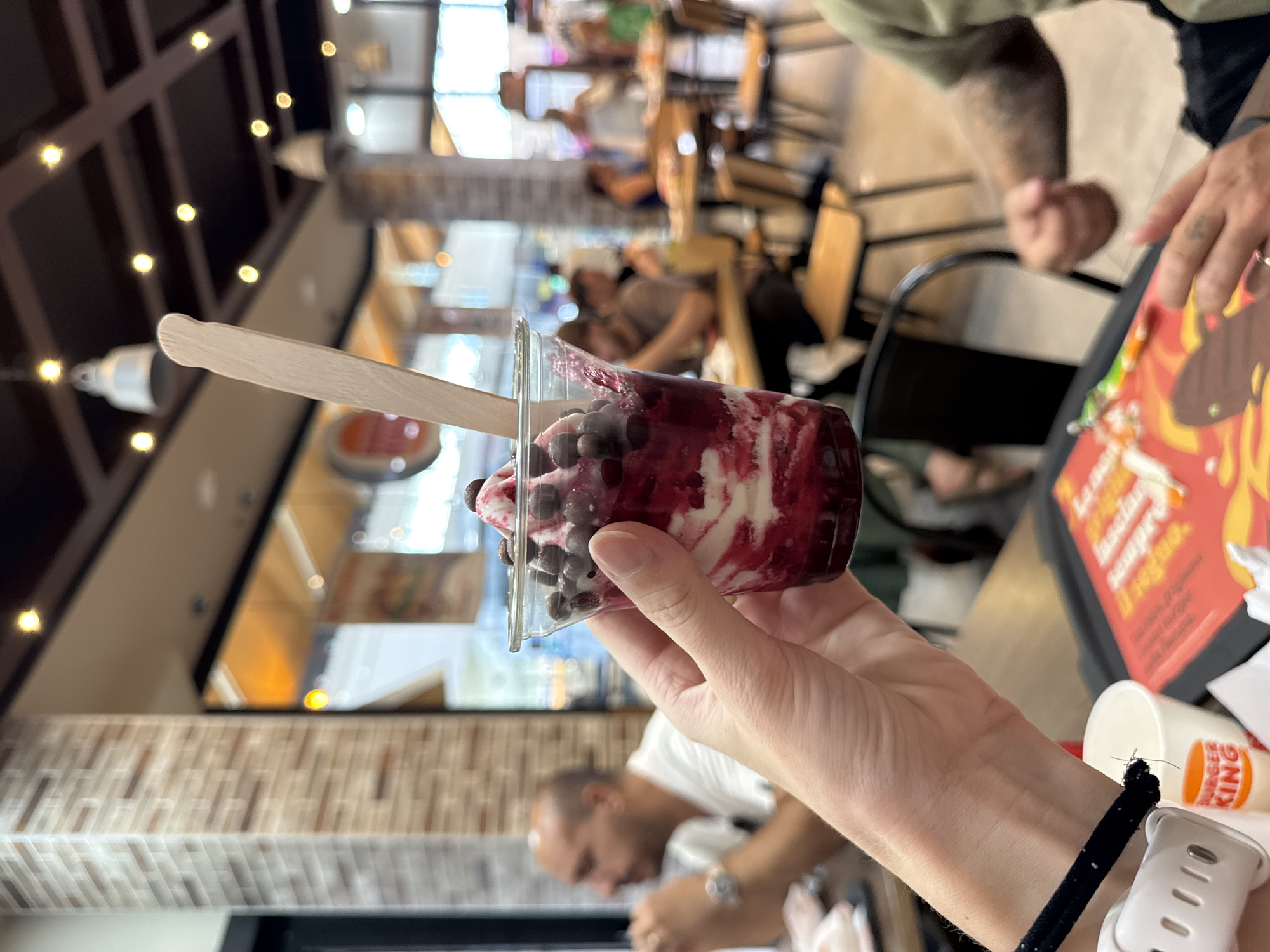 A person's hand holds a clear plastic cup filled with a red berry swirl soft-serve ice cream topped with dark chocolate chips and a wooden stick. In the background, several other people are seated at tables in what appears to be a Burger King restaurant. The lighting suggests it is daytime. The scene is indoors, likely a fast-food establishment.