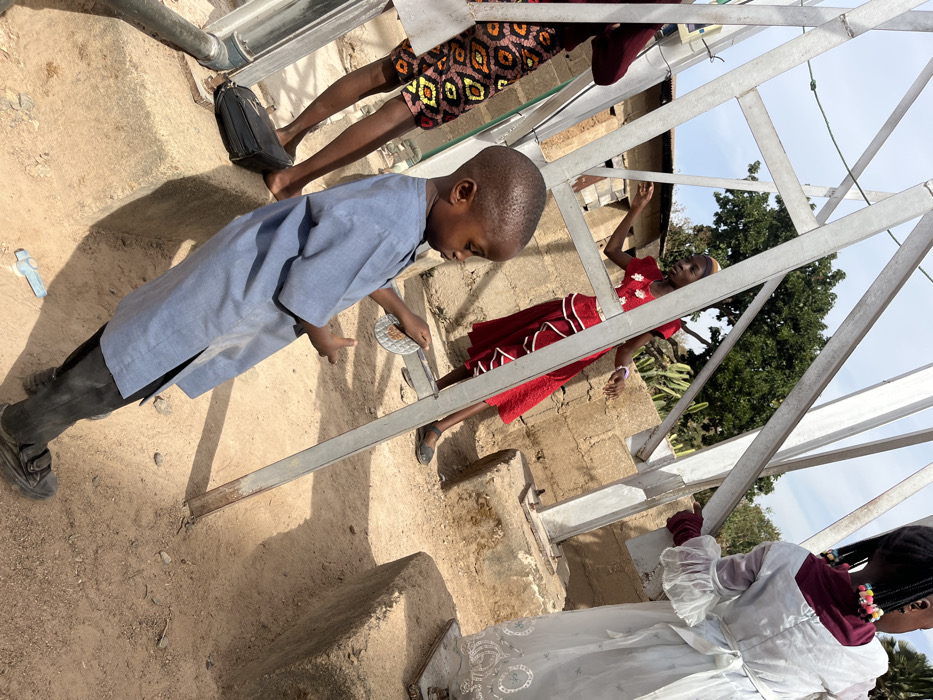 This image depicts an outdoor scene in Jos, Nigeria, featuring four individuals interacting with and around an unpainted metallic structural frame under strong sunlight. In the immediate foreground, a young boy, estimated 3-5 years old, wears a light blue tunic and black shoes with Velcro straps; he holds a small, round, reflective silver object in both hands, looking down at it. To his left, the lower half of an individual is visible, wearing a vibrant patterned skirt (brown, black, orange, yellow geometric design) and dark footwear, with a black shoulder bag positioned on the ground beside their feet. In the midground, a young female, possibly a teenager, wears a red dress with white tiered ruffles and white floral appliqués; she stands with arms slightly raised in a dynamic posture. To her right, partially obscured, the back of a young girl, approximately 5-8 years old, is visible. She wears a white long-sleeved dress with embroidered details on the hem and braided hair adorned with colorful beads.
The metallic structure consists of multiple diagonal and vertical silver-colored beams, anchored into rough concrete bases on the dry, sandy earth. A small, light blue plastic object lies on the ground near the young boy's feet. The background reveals unplastered concrete block walls and structures, a large green leafy tree, and spiky cactus plants, all beneath a clear blue sky. Distinct shadows are cast across the scene due to the bright sun.