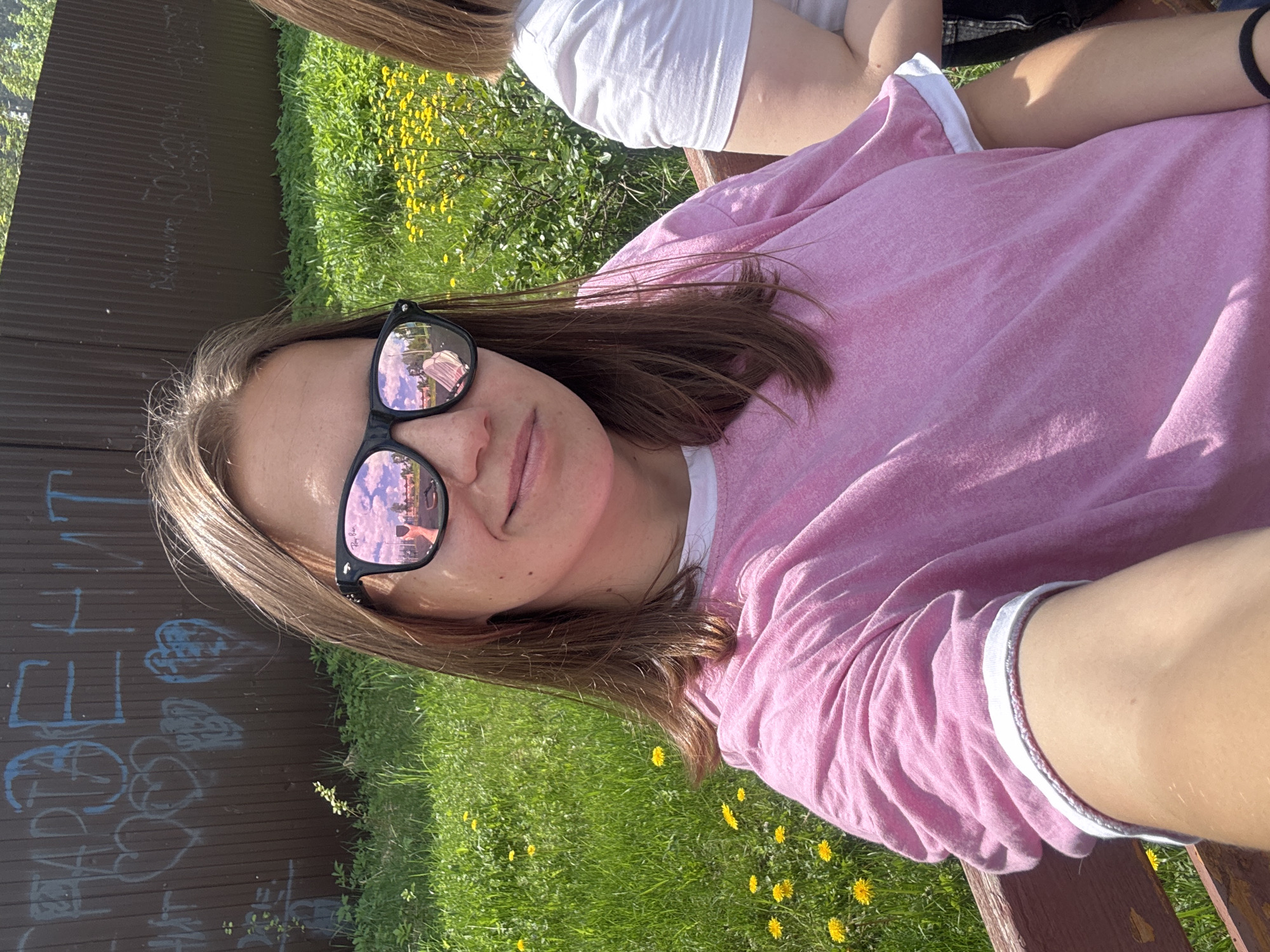 A woman wearing sunglasses and a pink and white shirt is sitting on a bench in Раздолье, Russia. The sunglasses reflect buildings and sky. A section of a brown fence with blue graffiti on it is visible on the left, along with green grass and yellow flowers. A second person wearing a white shirt can be seen partially in the top right corner.