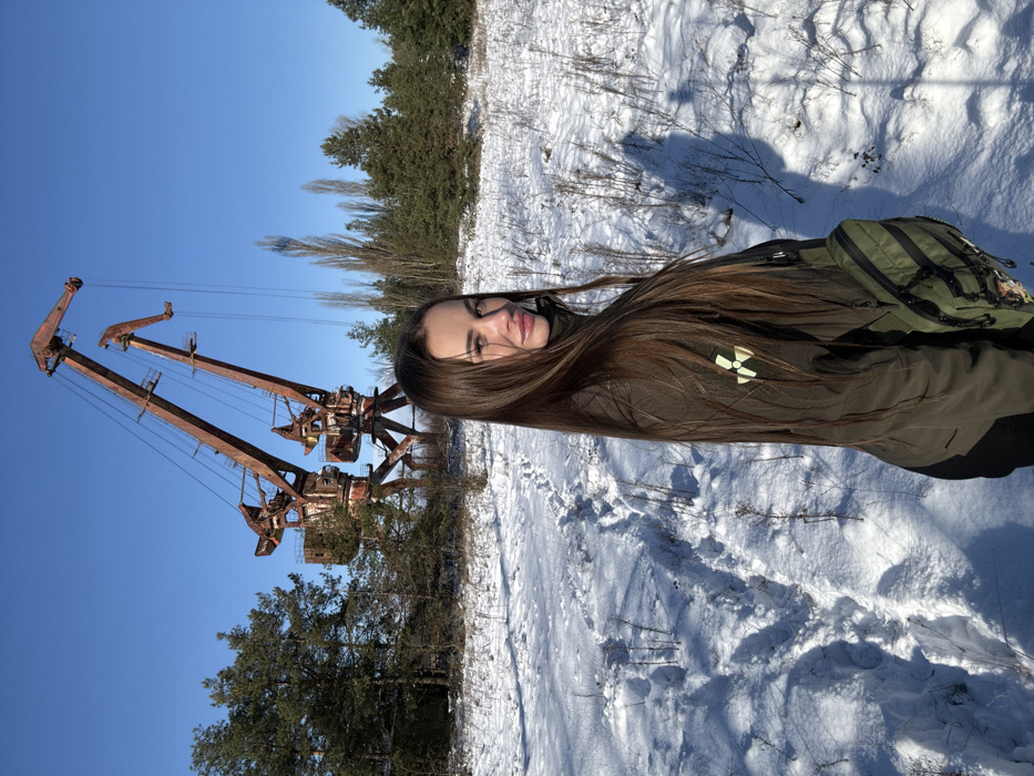 The image captures a winter scene at the Port of Prypiat, Ukraine. In the foreground, a young female individual stands partially facing away from the camera, looking over her right shoulder towards the viewer. She has long, dark brown hair and wears a dark olive-green jacket, which features a yellow radiation trefoil symbol on the left sleeve. A dark green tactical-style fanny pack or sling bag is worn across her front. The ground is covered in a layer of white snow, showing a distinct path of footprints leading away from the viewer towards the middle ground. In the background, against a clear blue sky, two large, heavily rusted, and disused gantry cranes, characteristic of the Port of Prypiat, dominate the skyline. Sparse coniferous trees and bare deciduous trees are visible in the surrounding snowy landscape.