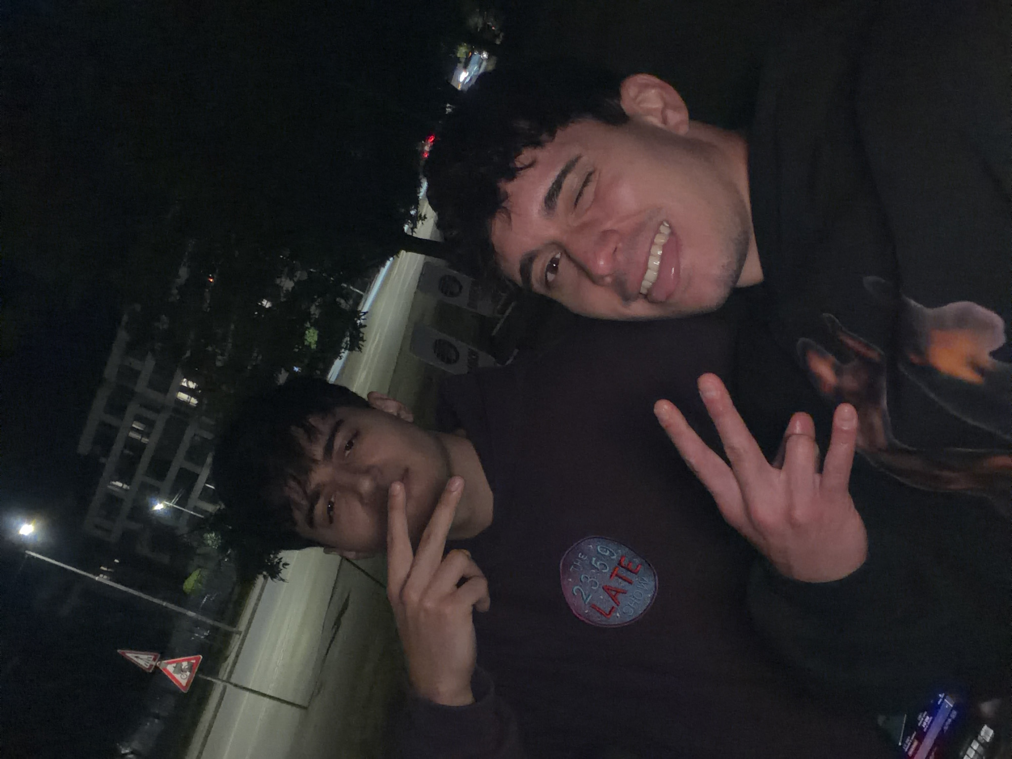 A tilted night-time photograph captures two young men in an urban setting, likely in Sofia, Bulgaria. The man in the foreground, positioned slightly left of center, wears a dark maroon long-sleeved shirt. A prominent circular patch on his shirt displays the text "THE 23:59 LATE SHOW". He faces the camera with a calm expression, making a peace sign (V-sign) with his left hand.
Behind him, closer to the right side of the frame, is another man with dark, curly hair. He grins widely, winking with his left eye, and holds up three fingers on his right hand. Both subjects convey a happy and casual demeanor.
The background is dark but illuminated by artificial light sources, including streetlights. Dimly visible details include multi-story buildings, silhouetted trees, and a paved road or sidewalk. Two triangular warning signs are visible on a pole near the road, one appearing to be a give-way sign and the other depicting a bicycle. The overall scene suggests a casual outing among friends in a city environment after dark.