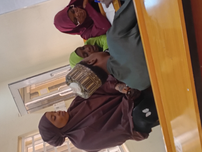 An indoor scene in Kwazailewa, Nigeria, shows four individuals seated at light-colored wooden desks. In the foreground, a male individual, viewed partially from behind, wears a dark green long-sleeved garment and a patterned cap; he holds a pen poised over a notebook. To his immediate left, a female wearing a dark brown hijab and matching attire is seated, observing the group; visible tattoos are present on her left forearm, and she wears black leggings with a white butterfly motif on the left leg. Behind the foreground male, a second female in a light green hijab is partially visible. To the right of the foreground male, a third female, wearing a dark red hijab and glasses, is focused downwards on written material. The background consists of light-colored walls, a barred window on the left, and a glass-paned door on the right.