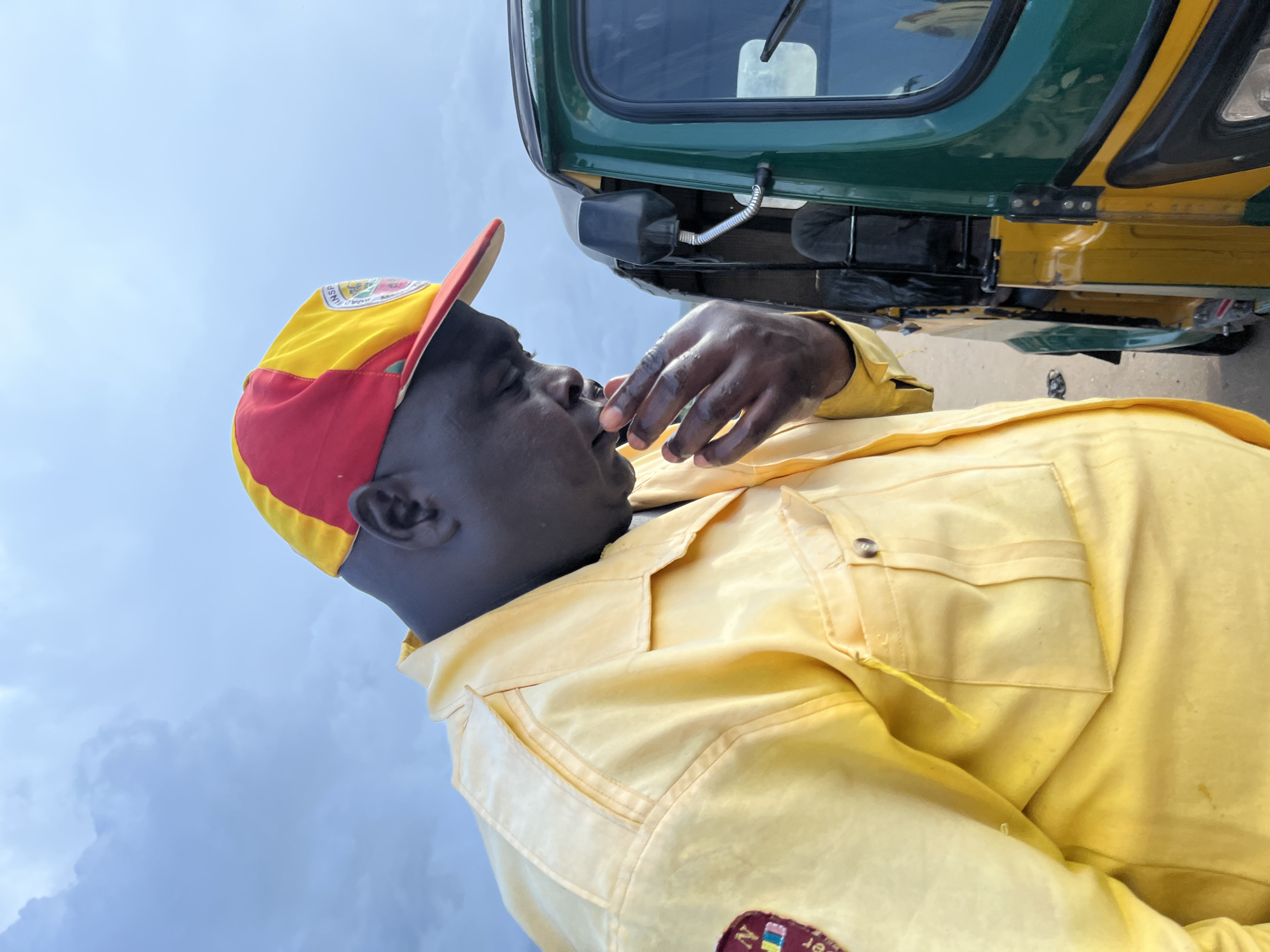 A man wearing a yellow shirt with red and yellow striped cap is looking down at his hands. He is standing next to a green and yellow auto rickshaw. The sky is cloudy and the scene is likely in a rural area of Nigeria, perhaps in the town of Rafin Par.
