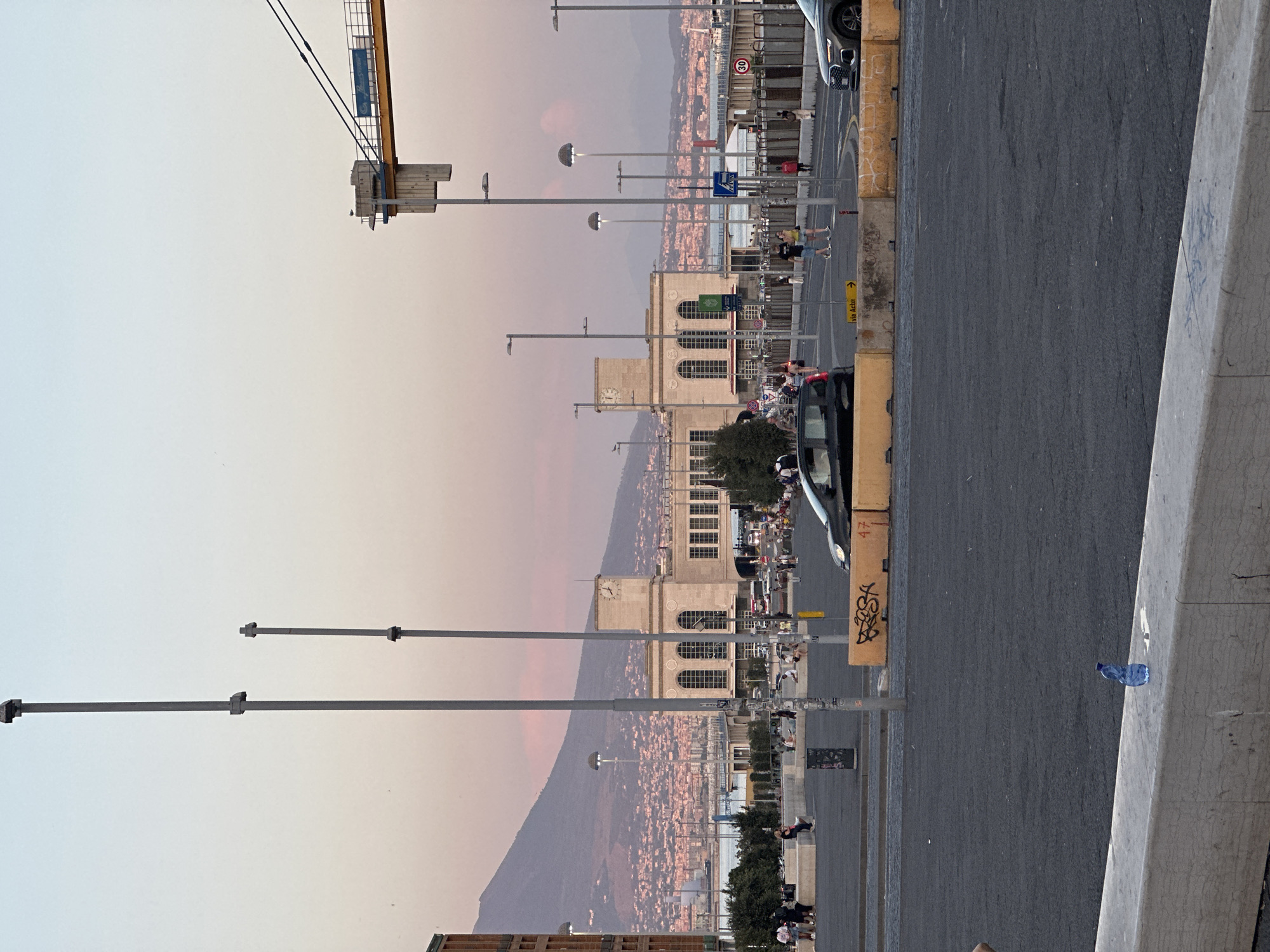 The image depicts a wide-angle, eye-level view of a public space in Naples, Italy, likely near the waterfront or a transportation hub, at dusk. In the background, Mount Vesuvius looms, its slopes dotted with the lights of the city. A large, classical-style building with arched windows and a prominent clock tower dominates the middle ground. Several streetlights with unique dome-shaped fixtures are visible throughout the scene. A yellow construction crane is partially visible in the upper right corner.
The foreground consists of a paved area with several concrete barriers, some marked with graffiti. A blue plastic water bottle sits on one of these barriers. People are scattered throughout the scene, some walking, others sitting on benches. Two cars are present in the mid-ground, one black and one dark gray. The sky exhibits a soft gradient of pale blue and pink, indicative of sunset. The overall impression is one of a busy urban environment transitioning into evening.