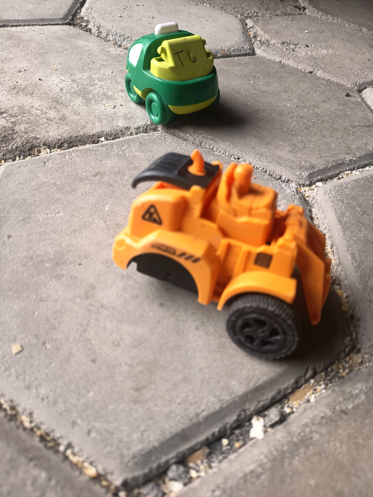 The image shows two toy vehicles on a paved surface in Sidorejo, Indonesia. In the foreground, an orange toy construction vehicle with black accents and a black wheel is visible. The vehicle has a hazard symbol on its side. In the background, a small green and yellow toy car is positioned slightly to the left. The pavement is composed of hexagonal gray concrete blocks, with gaps filled with sand and small stones. The lighting suggests it is daytime, with shadows cast by the objects. There are no people present in the image.