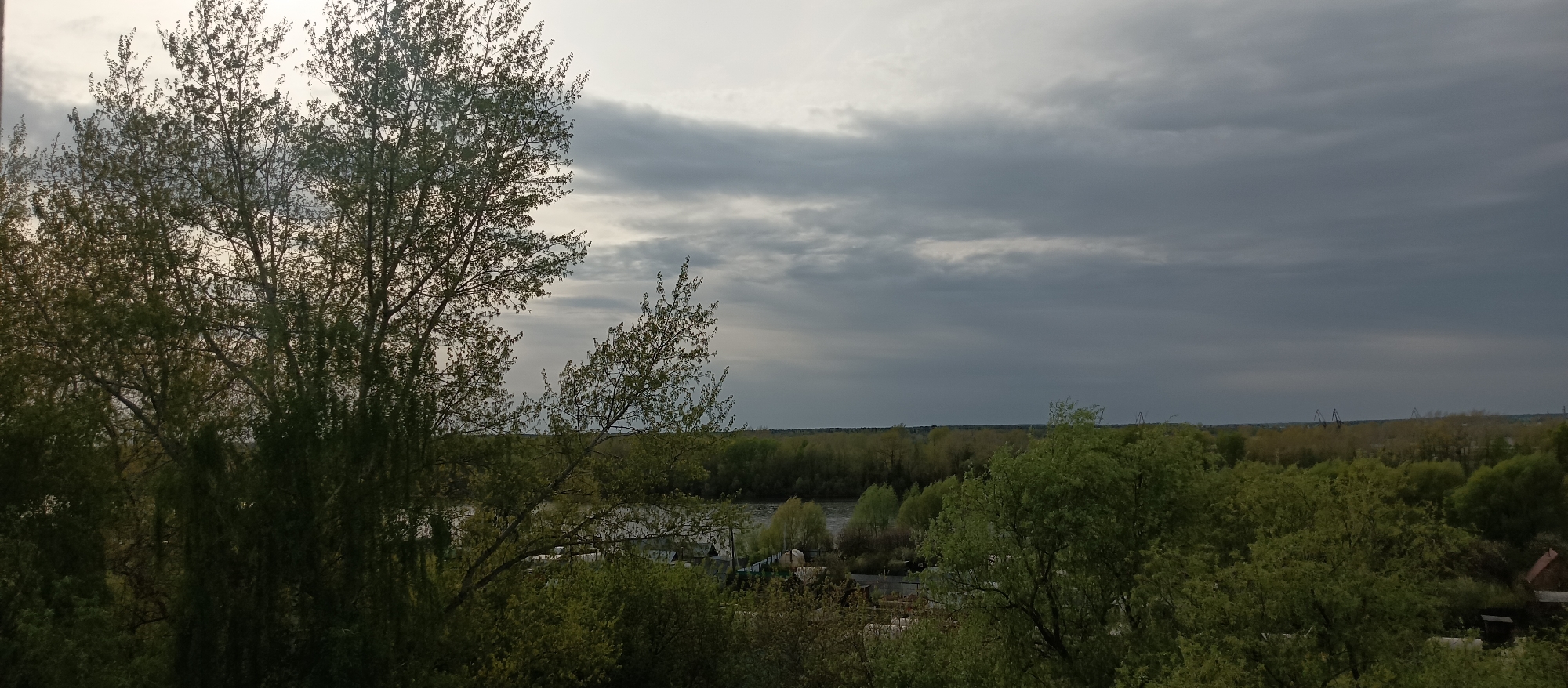 The image depicts a landscape view, likely from an elevated position, overlooking a river and a dense area of trees. The sky is overcast with heavy, grey clouds, suggesting potentially inclement weather. In the foreground, the focus is on a large tree with many branches and leaves, obscuring some of the view. The midground reveals a body of water, possibly a river, flanked by greenery. Beyond the trees and the river, there are distant structures, possibly industrial cranes. There are houses between the trees, surrounded by plants. The environment appears to be urban or suburban. Location: Unknown, Unknown.