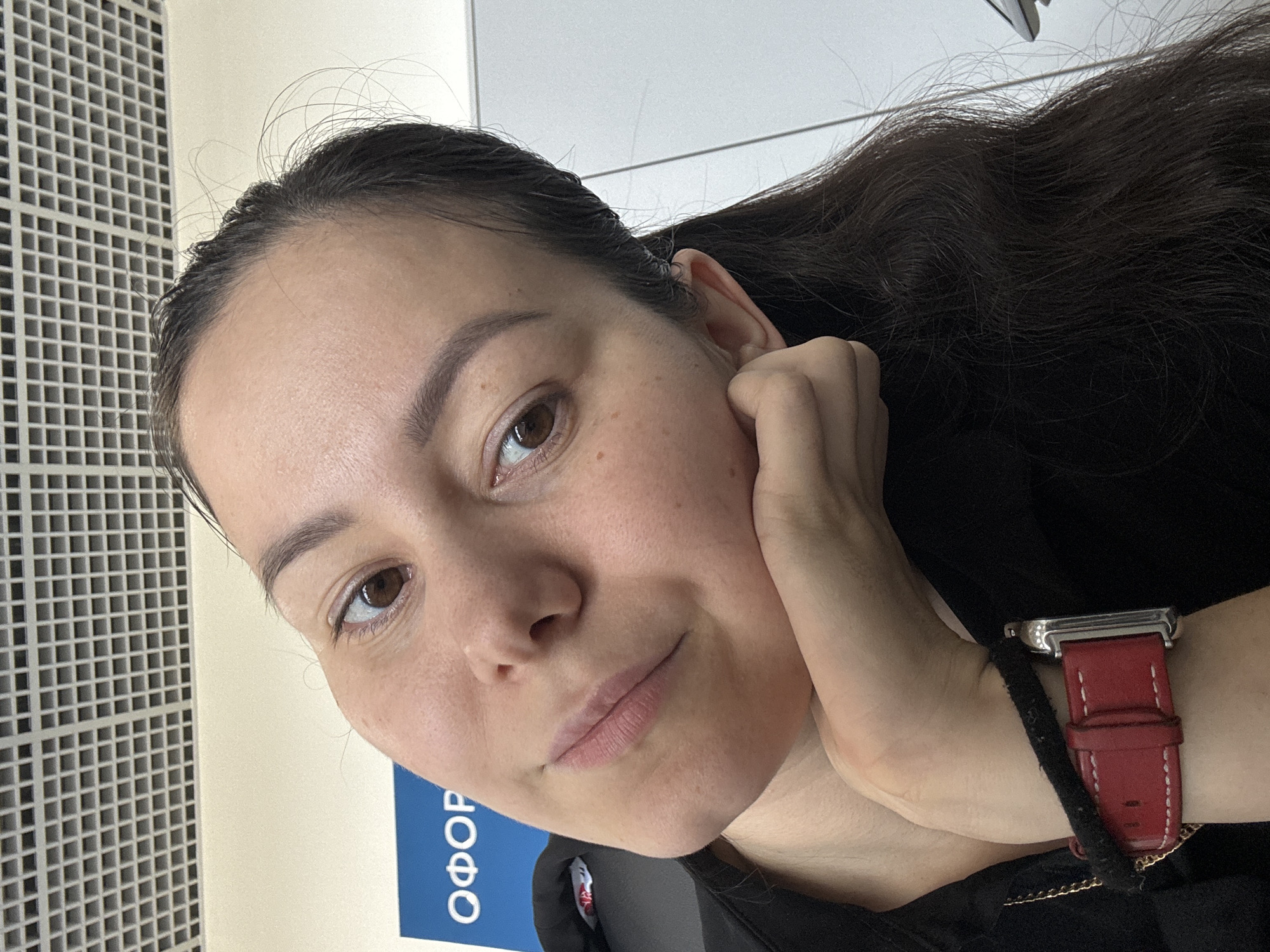 A woman with dark hair and brown eyes rests her head on her hand, looking towards the camera. She wears a black shirt and a red watch with a silver casing on her left wrist. Behind her, a blue sign with white lettering, appearing to read "ОФОР", is visible against a light-colored wall. The ceiling above is a grid of white acoustic tiles. The scene is indoors, likely in an office or public building in Vavilovo, Russia.