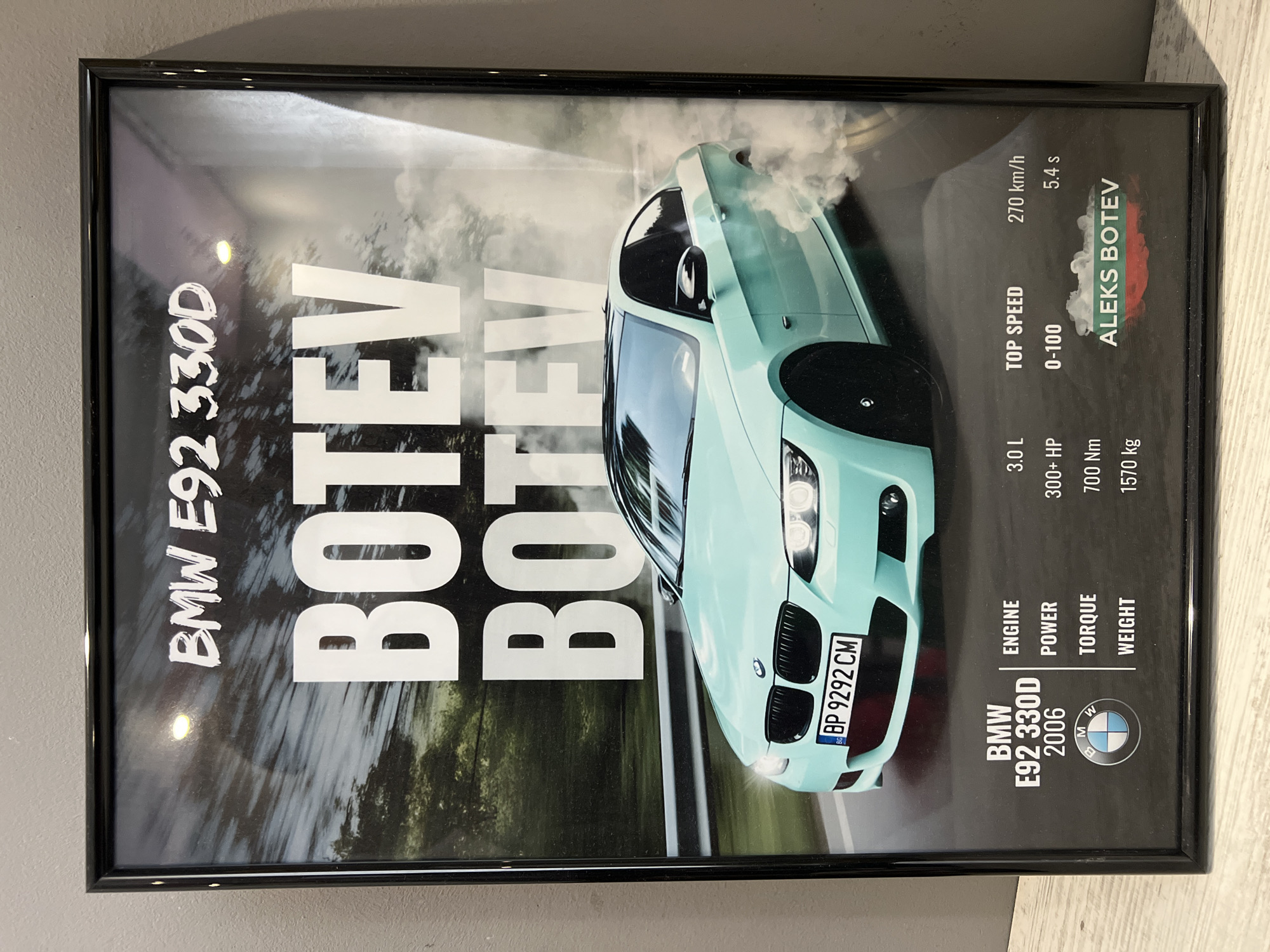 A framed poster displays a light blue (or mint green) BMW E92 330D coupe in a dynamic, performance-oriented shot. The car is centered on the poster, angled towards the viewer, with smoke or mist emanating from its front section, suggesting high speed or a burnout. The background is blurred, depicting a natural environment with trees and foliage, indicative of a road or track.
Visible text on the poster highlights the car's details and performance specifications. Large white text reads "BMW E92 330D" and "BOTEV" (repeated three times). The car's license plate, "BP 9292 CM," is visible, with "BP" indicating Plovdiv, Bulgaria, and a "BG" sticker confirming the country. To the right of the car, detailed specifications are listed: "ENGINE: 3.0 L," "POWER: 300+ HP," "TORQUE: 700 Nm," "WEIGHT: 1570 kg," "TOP SPEED: 270 km/h," and "0-100: 5.4 s." The name "ALEKS BOTEV" is also present, overlaid on a design resembling the Bulgarian flag. Below the specifications, a smaller "BMW E92 330D 2006" is listed alongside the BMW emblem.
The image itself is framed in black, and reflections on the frame suggest it is displayed indoors, under artificial lighting. The overall impression is a vibrant, enthusiast-focused presentation of a high-performance vehicle.