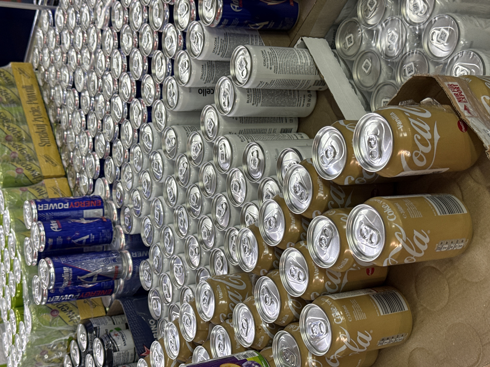 The image depicts a variety of canned beverages stacked on a shelf, likely in a retail setting. Brands visible include "Coca-Cola Vanilla", "Adrenaline Rush Energy Power", "Schweppes", "Stalcom", and another brand partially visible. The image is oriented with the cans arranged in rows, predominantly showing the top of the cans.