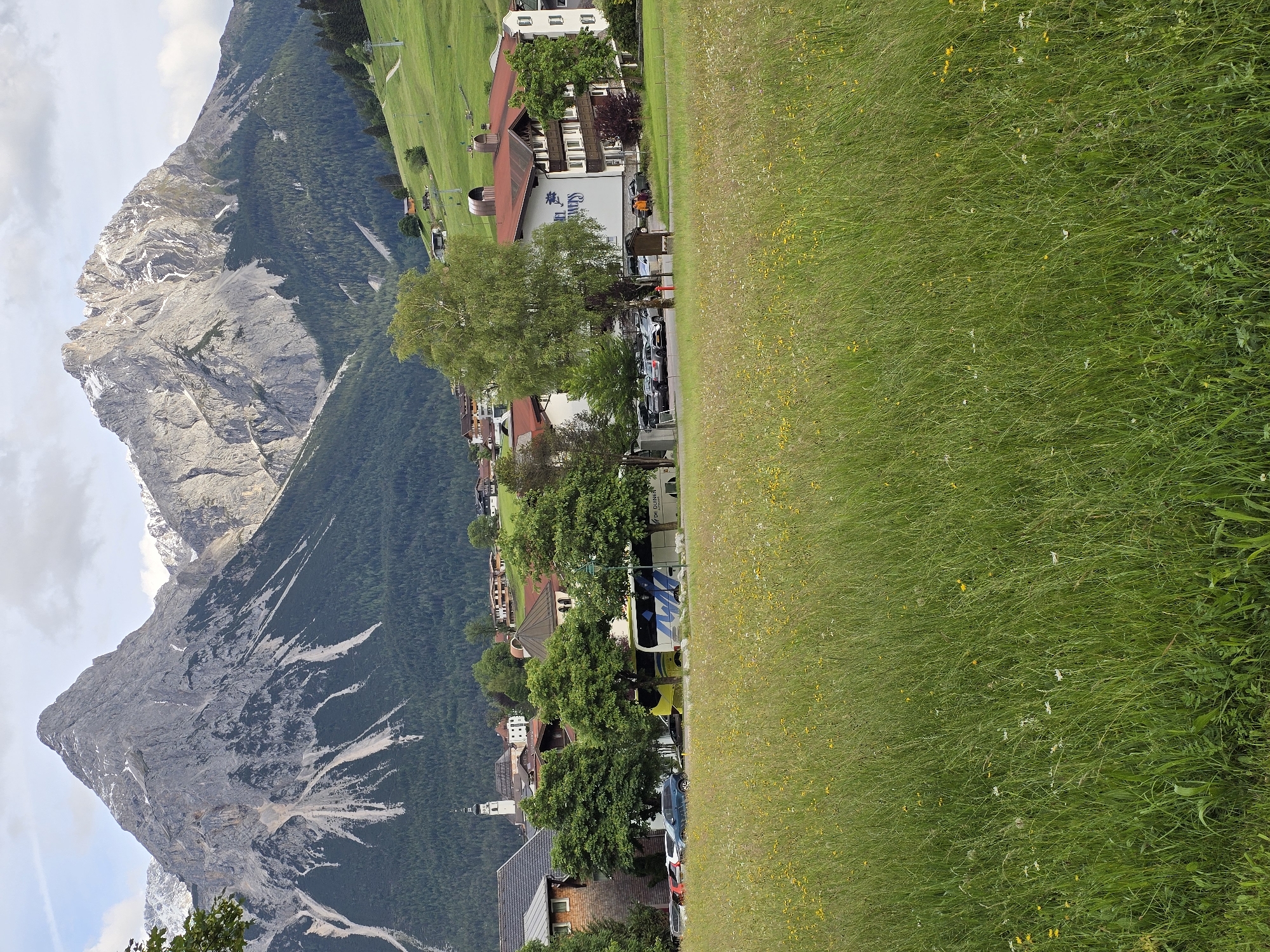 This is a vertical, eye-level, medium shot photograph of the village of Lermoos, Austria, situated at the base of a mountain range. The foreground is dominated by a field of green grass with scattered yellow and white wildflowers. Behind the field, a road runs horizontally across the frame, with several cars parked on the left. To the right of the road, a white and blue tour bus is parked, with the lettering "DE QUINCEY" visible on its side. Further to the right, a multi-story building with a red-tiled roof, identified as "Hotel Edelweiss" by its signage, stands prominently. Several trees, some with green foliage, are interspersed throughout the middle ground.
Beyond the village structures, the landscape rises into a densely forested mountainside. The background is defined by two imposing, jagged mountain peaks. The left peak is gray and rocky with white snow patches near its summit, and steep, eroded gullies descending its face. The right peak is also rocky and snow-capped, with dramatic, rugged terrain. The sky above is partly cloudy with patches of blue. A church steeple is visible in the village on the far left. The overall scene depicts a tranquil alpine village nestled within a dramatic mountain landscape.