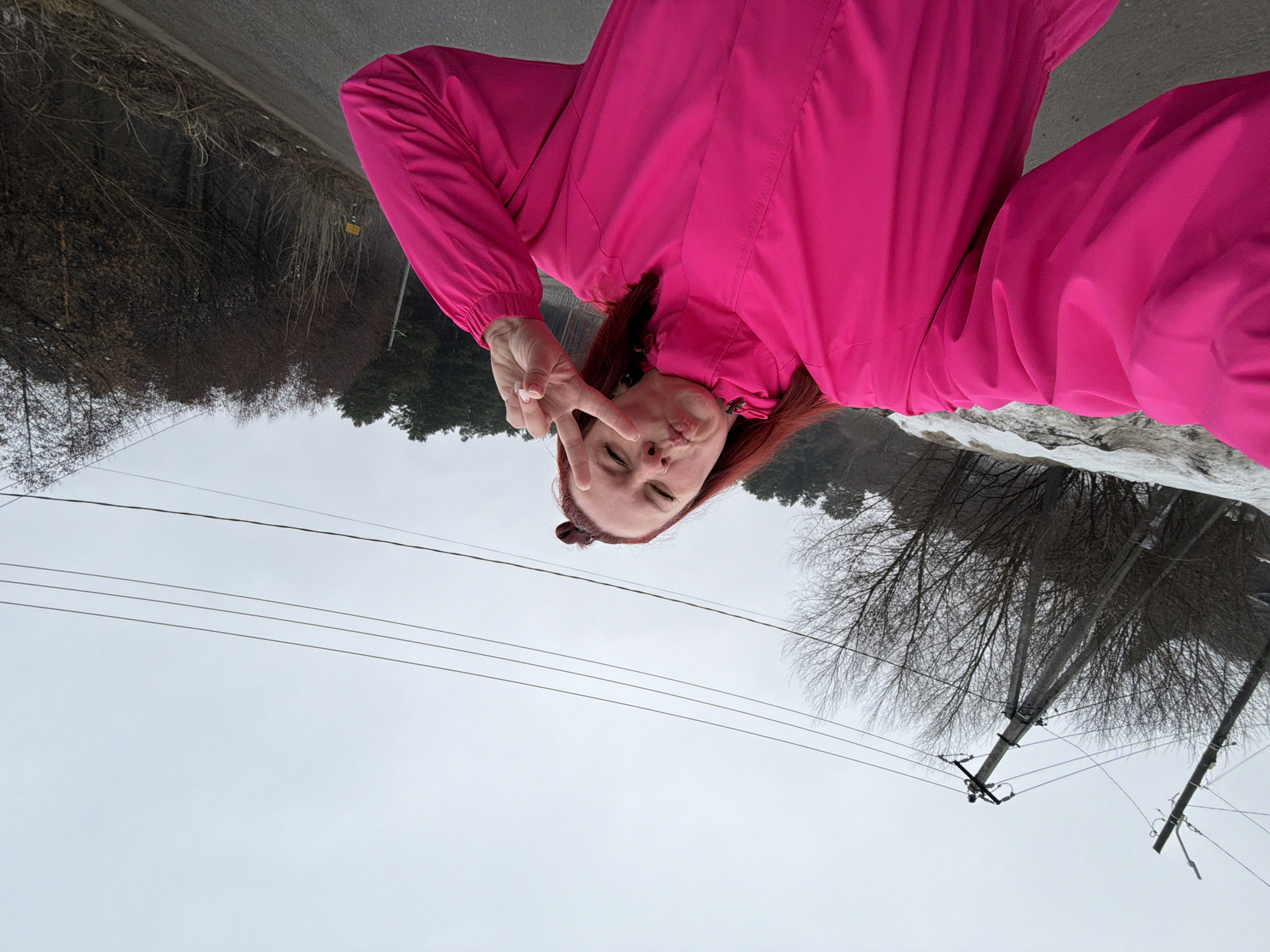 A woman in a bright pink snowsuit poses for a selfie, holding up a peace sign near her eye. The location is near the city of Chaikovsky, Russia, evidenced by the presence of snow, bare trees, and utility poles with overhead power lines. A road is visible in the background.
