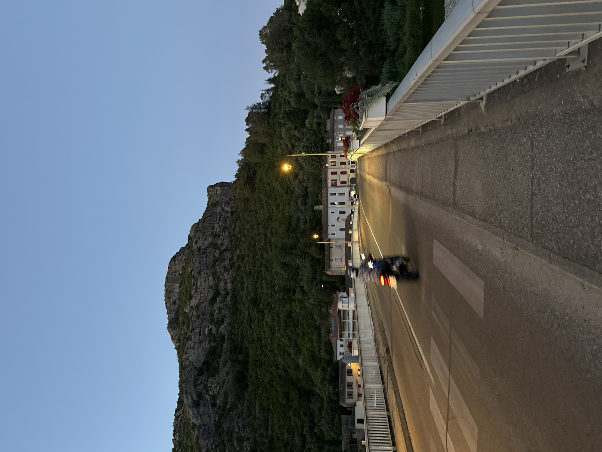 The image shows a bridge over a river in Anduze, France at dusk. A motorcycle is visible on the bridge, with a rider on it. The bridge has a pedestrian walkway and a road with painted lane markings and a crosswalk. Buildings are visible on the left side of the bridge, and a hill covered in trees is in the background. A street lamp is lit on the right side of the bridge. The sky is clear and a pale blue.