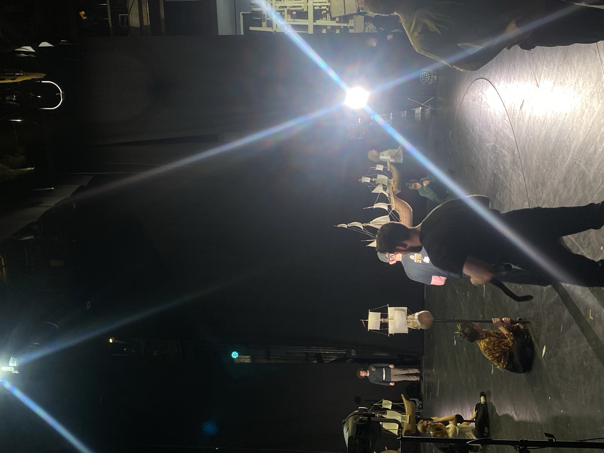 The scene appears to be a stage setup, possibly for a play or performance. There are several people present, some of whom appear to be actors or performers. One person is holding a prop resembling a ship. Other props and equipment are visible. The environment appears to be a dark stage with lighting equipment. The location, Rusanovo, Russia, cannot be determined from the image.