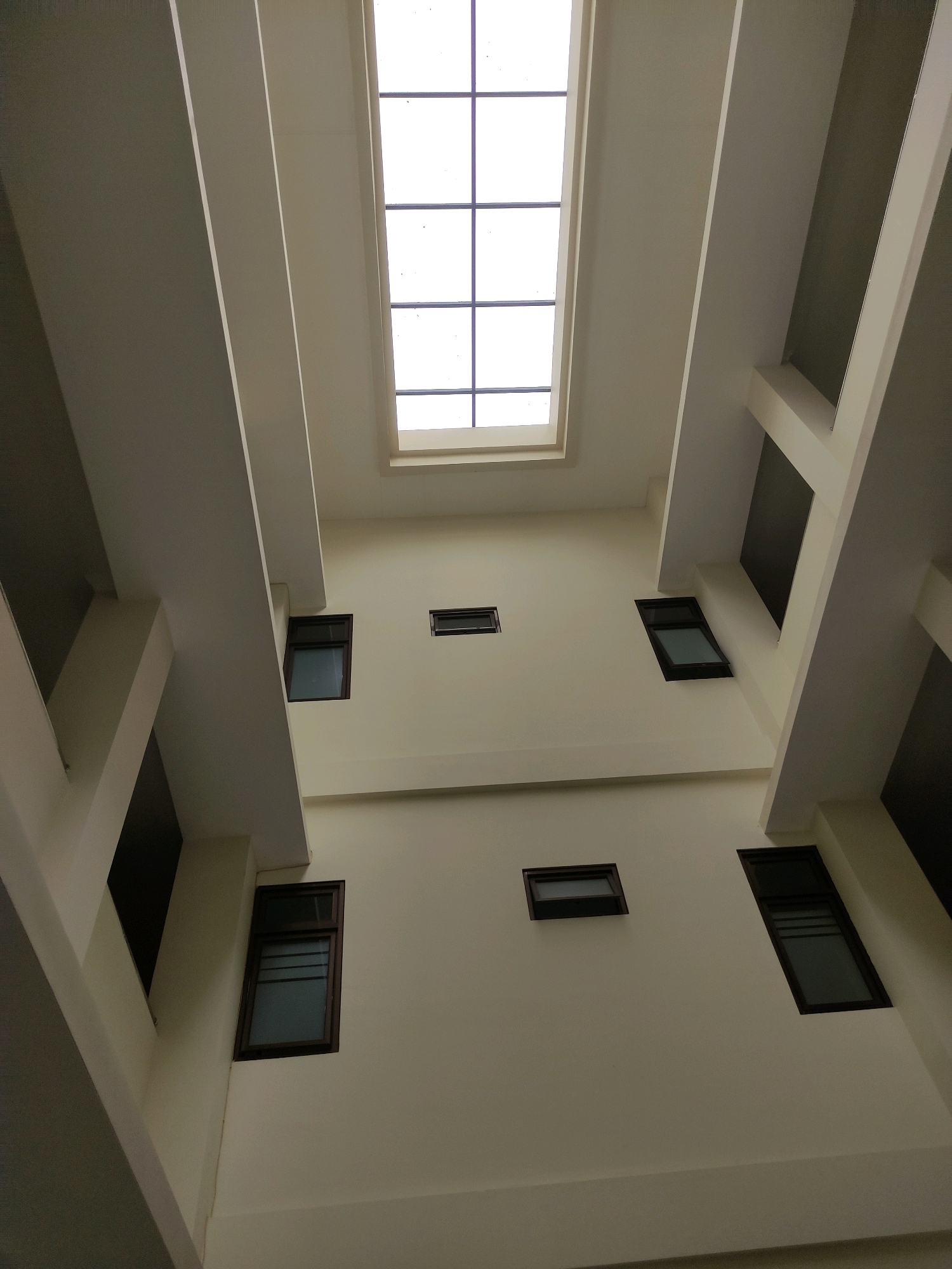 This image depicts the interior atrium of a modern building, likely an educational institution or office complex, with a focus on the architectural interplay of light and shadow. The viewpoint is looking upwards, towards a large, rectangular skylight at the apex of the structure. The skylight is divided into a grid by dark mullions. Surrounding the atrium are multiple levels of balconies or walkways, indicated by thick, cream-colored beams that extend diagonally inwards. Numerous windows of varying sizes and orientations punctuate the cream-colored walls of the building. These windows are uniformly framed in dark brown or black, and their panes are tinted, obscuring any clear view of the interior spaces behind them. Some windows are open, revealing a darker interior, while others appear closed. The architectural design emphasizes geometric shapes and the sense of depth created by the receding levels and the central light source. The overall impression is one of a clean, contemporary, and well-lit interior space. This building is identified as the Atrium Gedung Layanan Disiplin Mahasiswa at Universitas Muhammadiyah Purworejo, located in Purworejo, Indonesia. No people are visible in the image.