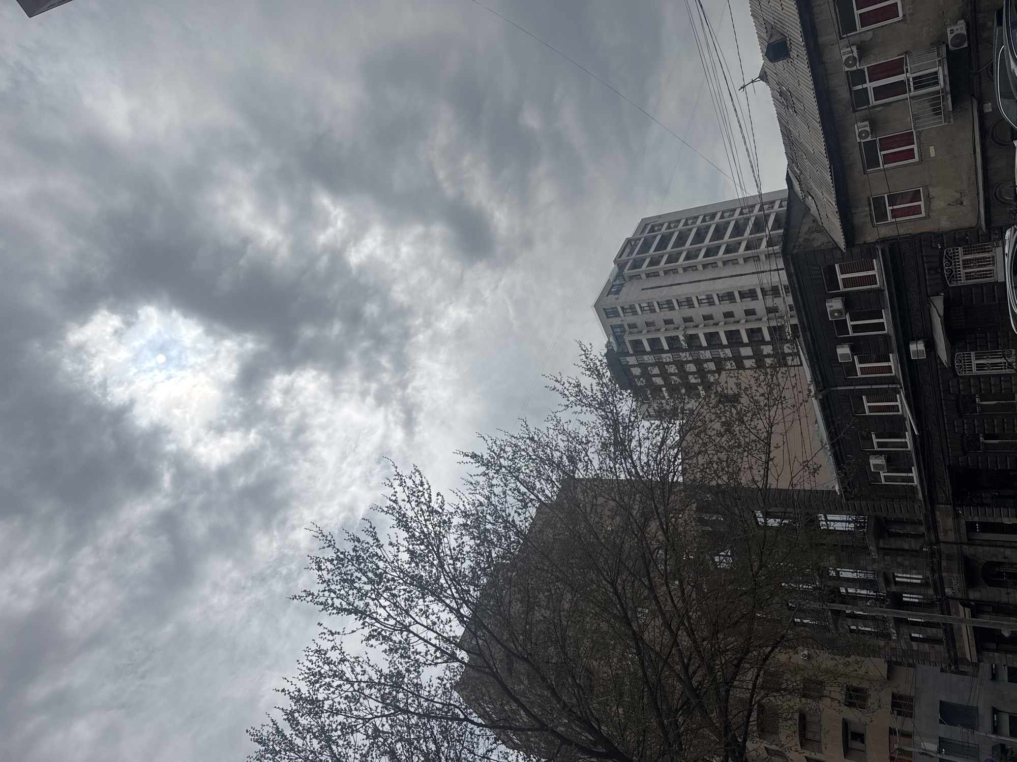The image shows a low-angle view in Yerevan, Armenia. A cloudy sky obscures the sun. Buildings of various heights and architectural styles are visible. A tree with bare branches occupies the lower portion of the frame, obscuring some of the buildings. Utility lines cross the upper portion of the image. Air conditioning units are mounted on the side of one building. There are no people visible in the frame.