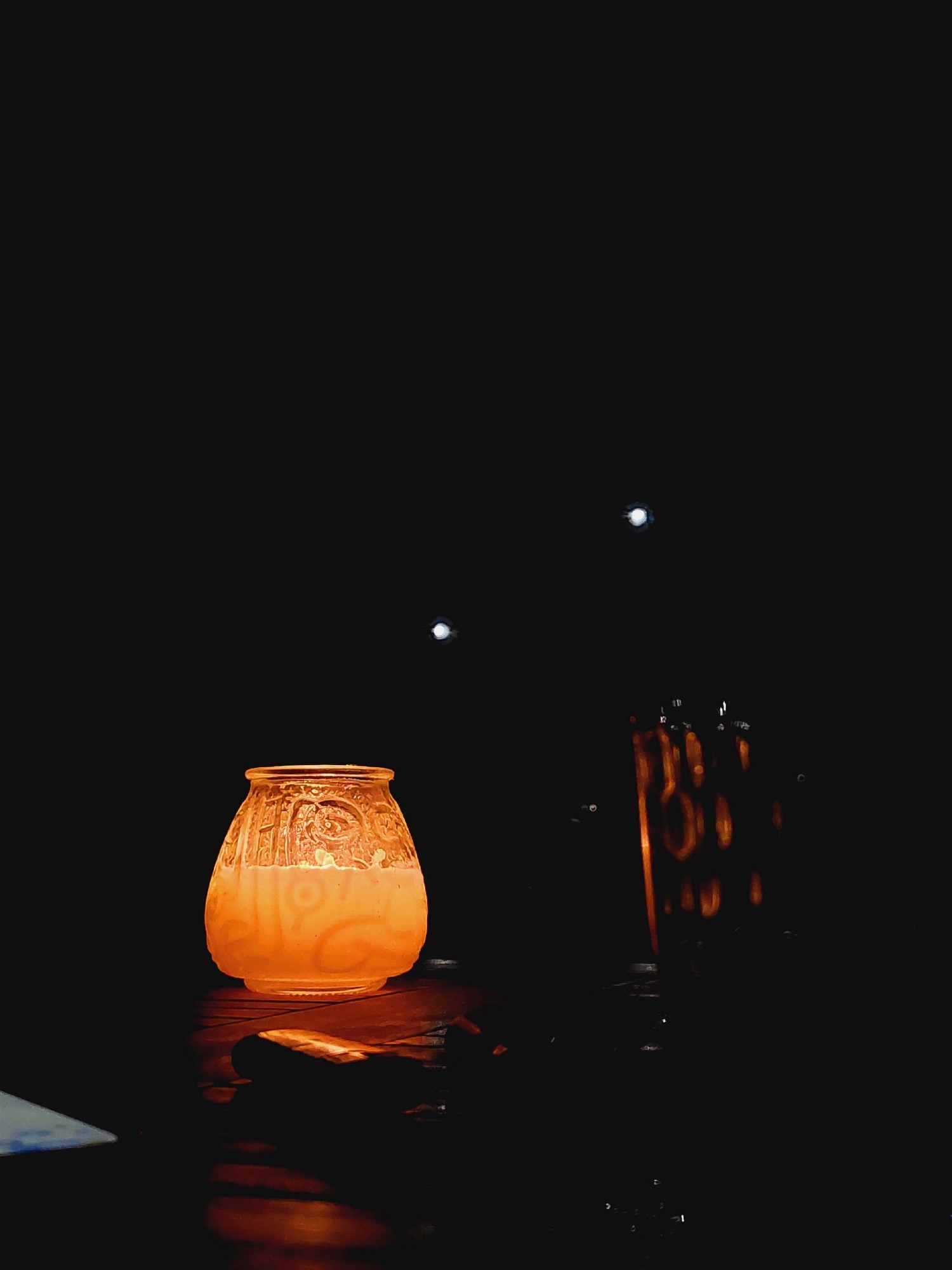A warmly lit, ornate glass lantern sits on a dark, reflective surface, casting an orange glow. In the background, two small, bright lights are visible against a black sky, possibly distant lights or stars. To the right of the lantern, a blurred, upright object with vertical, illuminated lines is also present, suggesting another light source or decorative element. The scene is dark and atmospheric, with the lantern being the primary illuminated subject. The location of Sozopol, Bulgaria is not visually represented in the image.