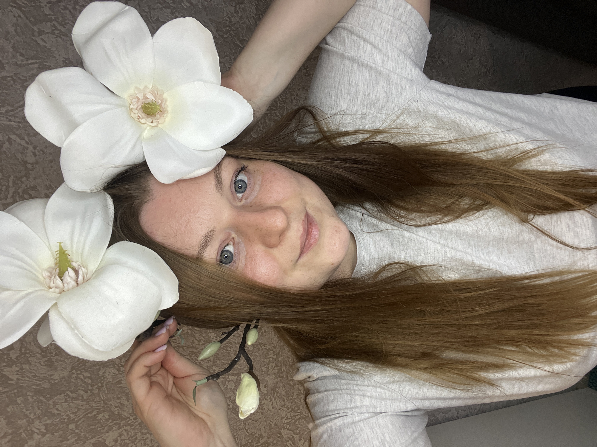 A woman with auburn hair and blue eyes is lying down, facing upward. Two large white magnolia flowers are placed around her head. She is wearing a light gray t-shirt. Her left hand is visible holding a small branch with buds. The background appears to be a textured surface, possibly a rug or carpet. The location of Раздолье, Russia is not evident in the image.