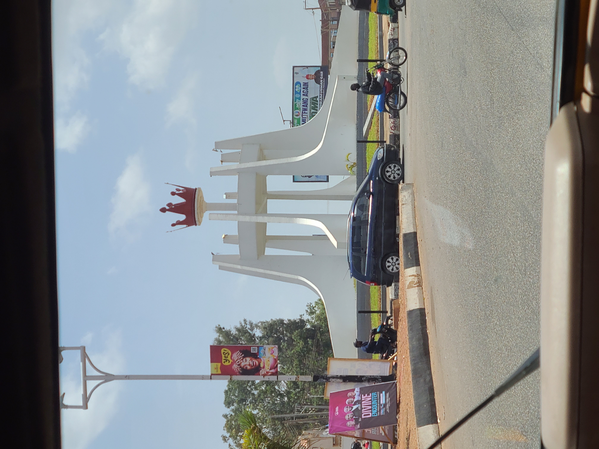 The image captures a scene in Barakin Lerawi, Nigeria, focusing on a prominent white monument topped with a red crown. Adjacent to the monument is a dark-colored car parked along the roadside. Nearby, two motorcycles are parked, one of which has a rider sitting on it.
Along the side of the road, advertising banners are visible, including a "Yes!" brand advertisement, and a banner promoting a "Divine Encounter" event. Overhead power lines crisscross the frame. The sky is light blue with scattered clouds. The photograph seems to have been taken from inside a vehicle.