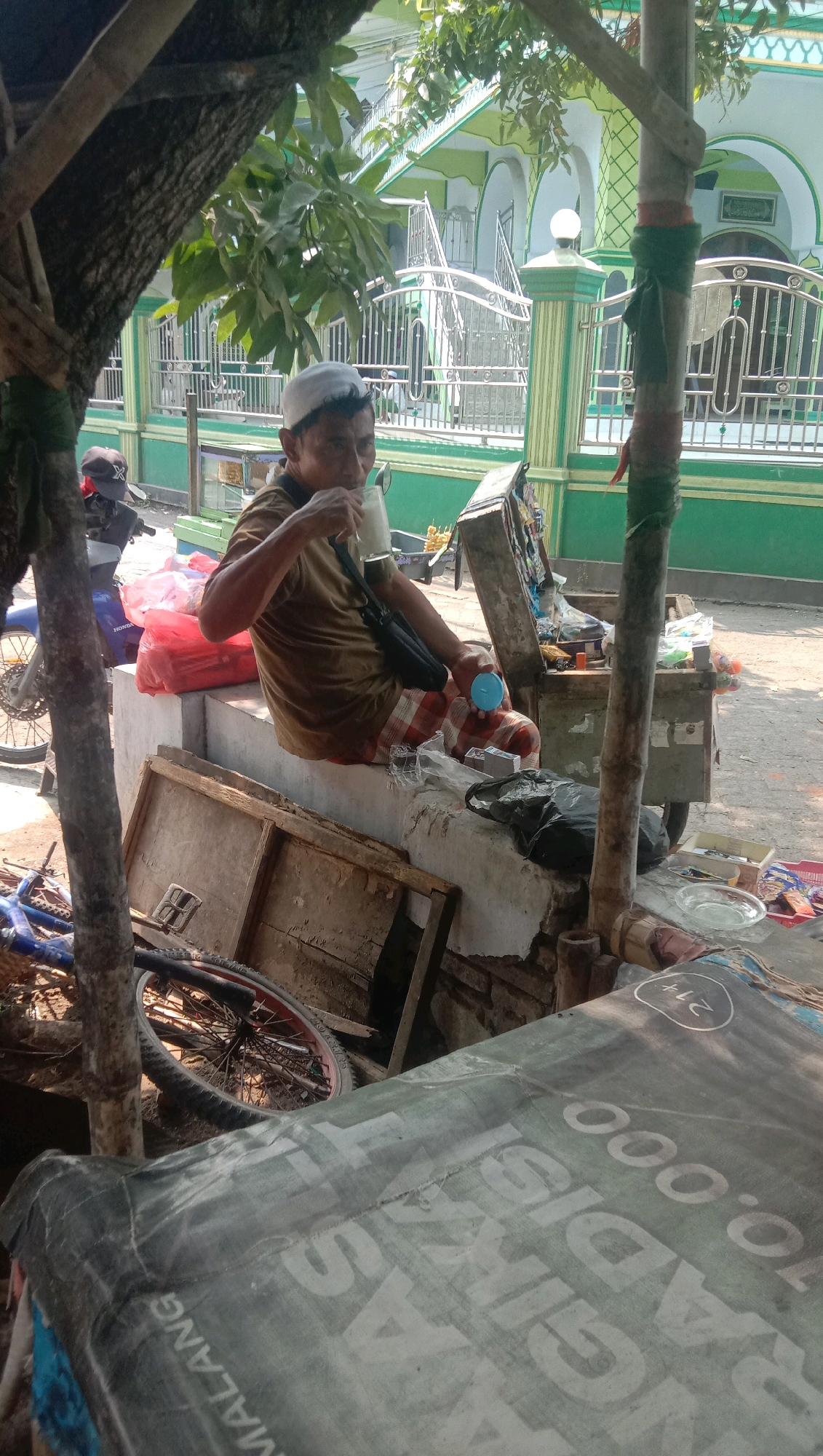 A man wearing a white cap and a brown shirt is sitting on a raised concrete platform, drinking from a glass and holding a blue cup in his other hand. He is in an outdoor setting with a building that has green and white elements in the background, suggestive of a mosque or religious building. A motorcycle is parked to his left, with red bags on the ground next to it. Various items are scattered around, including a wooden crate, a bicycle wheel, and a cart with goods. A large, patterned tarp is visible in the foreground. The overall impression is that of a street-side scene in a somewhat cluttered environment.
