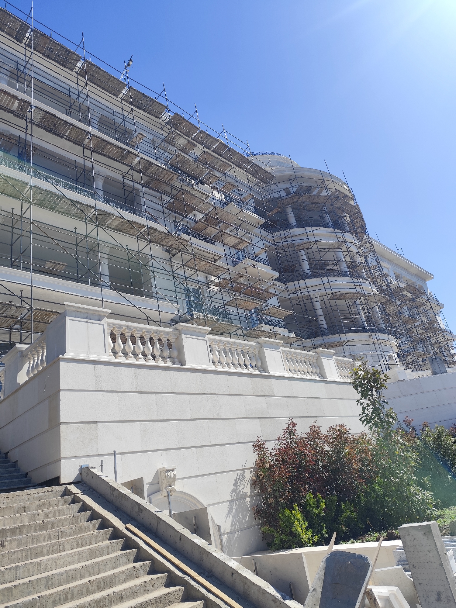 The image shows a large, multi-story building under construction or renovation, extensively covered in scaffolding. The building features white architectural details, including balconies and balustrades. A set of concrete stairs leads up to the building's raised platform. Vegetation, including shrubs and small trees, is present around the base of the building. The sky is clear and blue. The location is a city and country that are unknown.