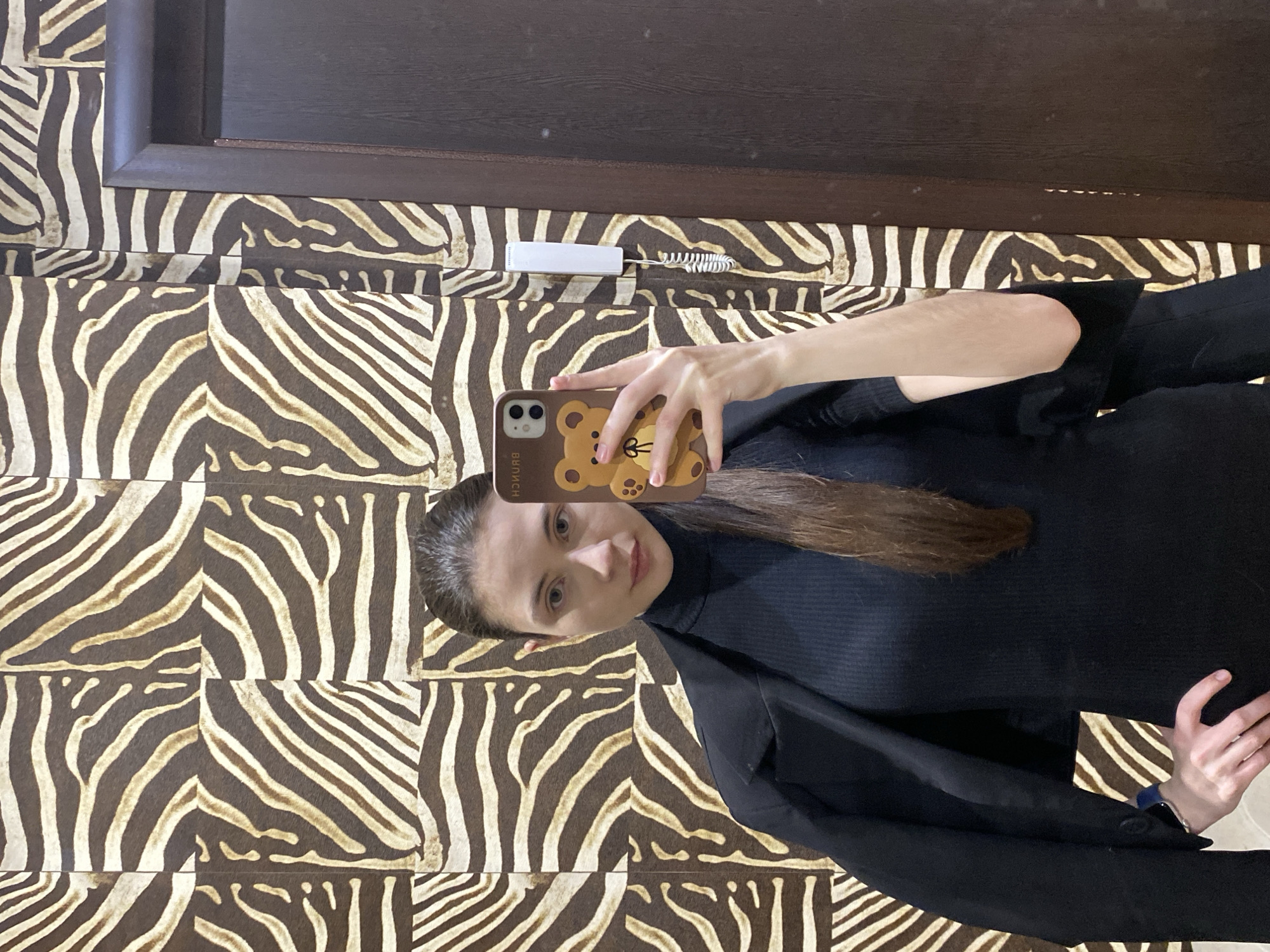 A woman is taking a selfie indoors, likely in a room with walls covered in zebra-patterned wallpaper or tiles. A dark brown door or panel is visible above the zebra pattern. The woman is holding a phone with a brown teddy bear case. A white device with a coiled cord is mounted on the wall near the top of the frame. The woman is wearing a black turtleneck sweater and a black jacket. The specific location in Votkinsk, Russia cannot be determined from this image.