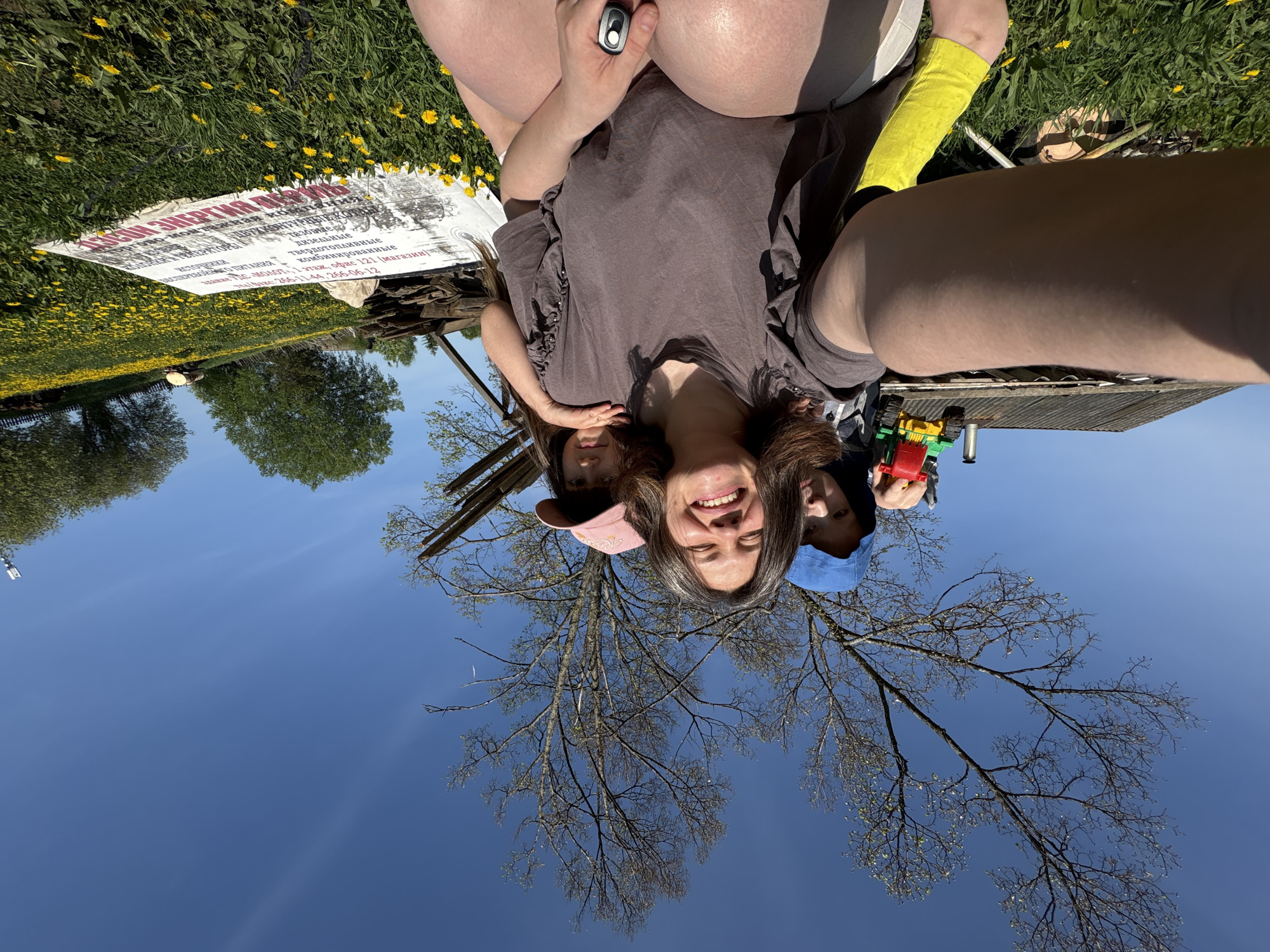 The image shows a selfie taken by a woman with two children. The background is an outdoor scene featuring a blue sky, a tree with bare branches, grass, and dandelions. Part of a dilapidated wooden structure is visible, along with a sign containing Russian text ("тазовые дизельные твердотопливные комбинированные Здание УЛС Молот, 1 этаж, офис 121 (магазин) Факс: 266-11-44, 266-06-12"). The text indicates a location in Russia (City Nizhnyaya Chermoda). The children appear to be a young girl in a pink cap and a young boy in a blue cap. The boy is holding a toy.