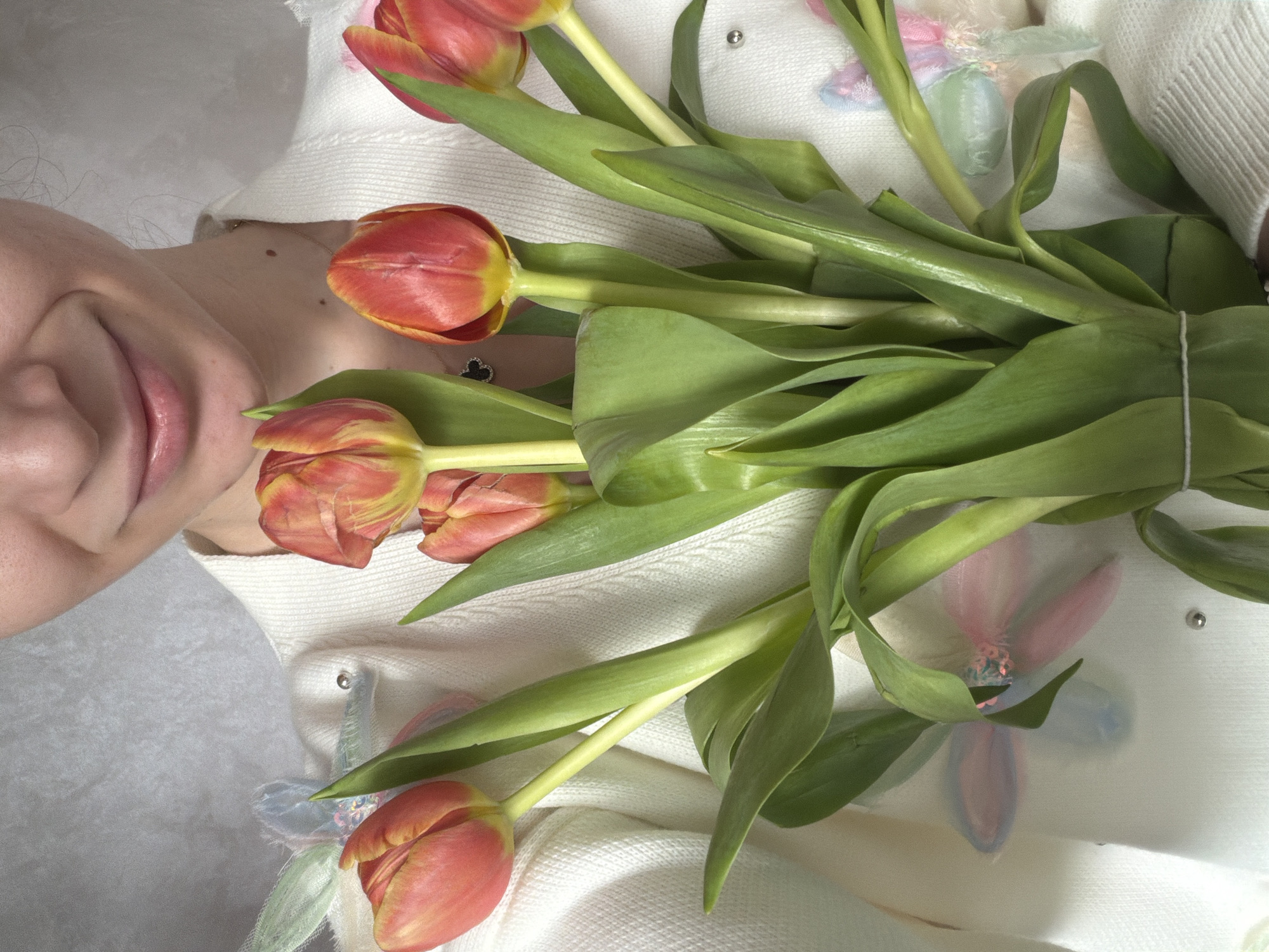 A close-up, high-angle shot depicts a woman lying down, her head visible in the top left corner. She is smiling slightly. A bouquet of red and yellow tulips rests on her chest, obscuring part of her white sweater. The flowers are tied together with a white string. The sweater appears to have floral embellishments. The background is a slightly textured, off-white fabric. The location and city are not discernible from the image.