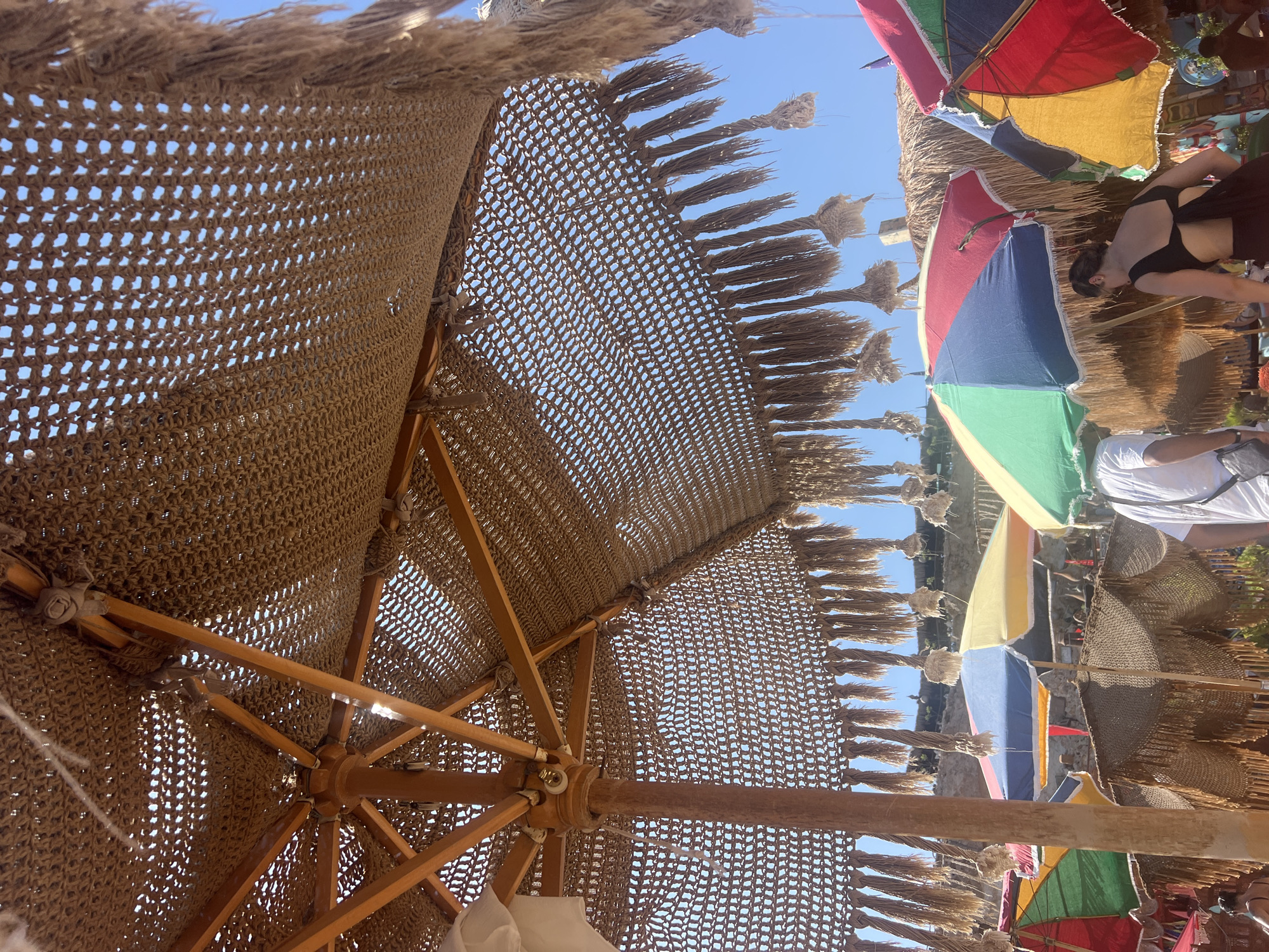 An outdoor scene under a bright blue sky at what appears to be a beach or resort area. The foreground is dominated by a large, open beach umbrella with a woven, straw-like canopy and fringe detailing, allowing sunlight to filter through. The umbrella's wooden frame and central pole are visible. Behind this, several other beach umbrellas are spread out, some with vibrant colored panels (red, yellow, blue, green) and others with similar woven canopies. In the midground, a person wearing a white t-shirt and dark pants is walking away from the camera. To the right, a person in a black top and dark bottoms is visible from behind, facing away and looking down. The background shows a sandy or rocky embankment with some structures and vegetation, suggesting a coastal environment. The overall atmosphere is casual and summery, characteristic of a vacation spot like Sharm El Sheikh, Egypt.