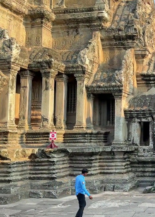The image shows a man in a blue long-sleeved shirt and black pants walking on the stone ground in front of a large, intricately carved stone structure, likely part of Angkor Wat in Siem Reap, Cambodia. The structure features multiple levels of tiered stonework, ornate columns, and what appear to be decorative carvings on its upper sections. A red and white sign with a circular symbol is placed on a raised stone platform to the left of the man. The lighting suggests it is either early morning or late afternoon, with warm sunlight illuminating the ancient architecture. The man is walking away from the camera, his posture suggesting he is moving at a casual pace. The overall scene depicts a historical landmark with a visitor present.