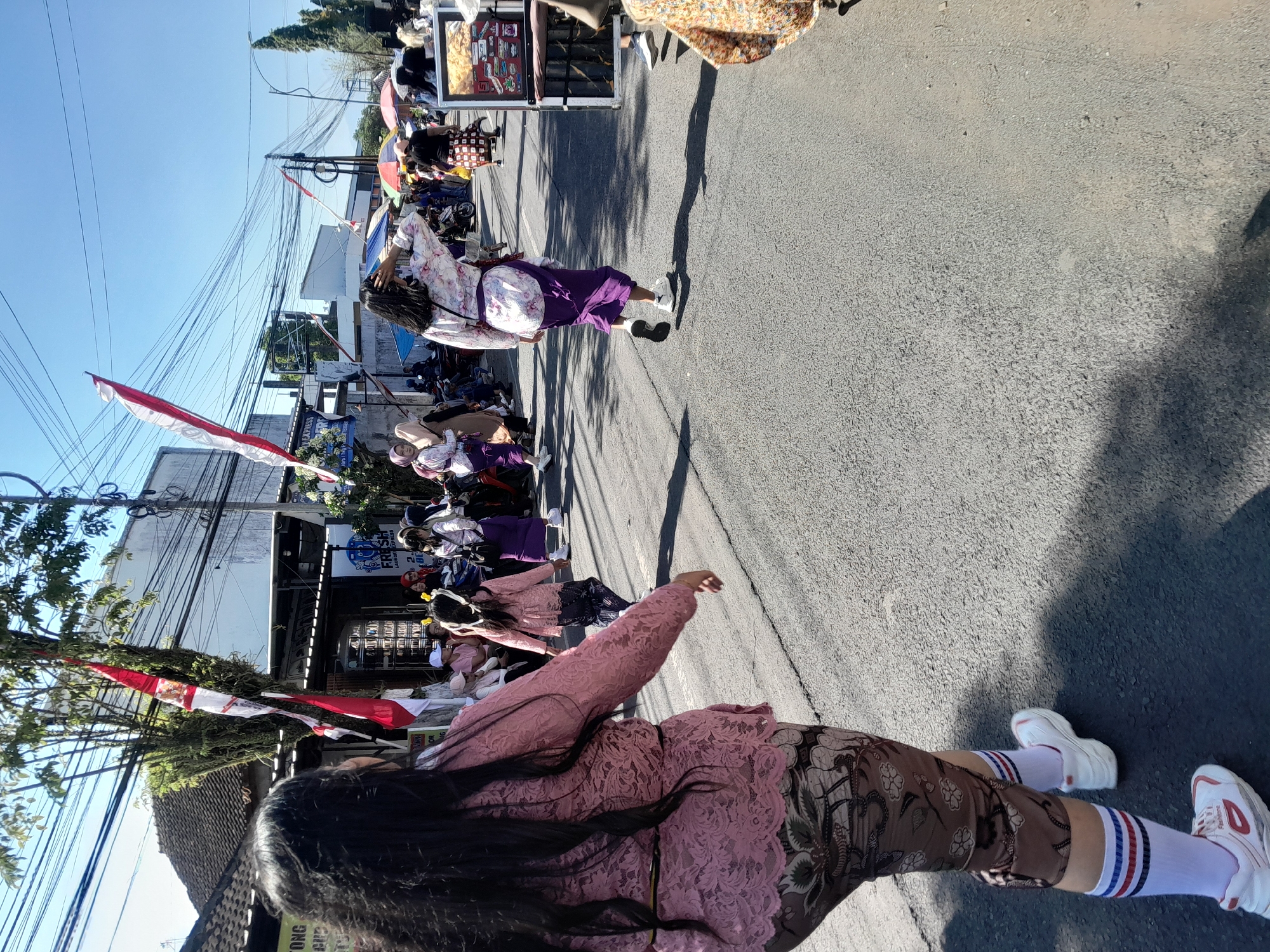 In Gentengwetan, Indonesia, a group of people are walking on a paved street under a clear blue sky. The image focuses on a woman in the foreground with long dark hair, wearing a pink lace top and patterned leggings with white socks and sneakers. She is walking away from the camera, to the left of the frame. Behind her, other women are seen in similar traditional attire, some carrying accessories or wearing headscarves. Red and white flags are displayed on poles along the street, suggesting a celebration or event. In the background, buildings, power lines, and parked motorcycles are visible. The ground is asphalt, with shadows cast by the people and surrounding structures.