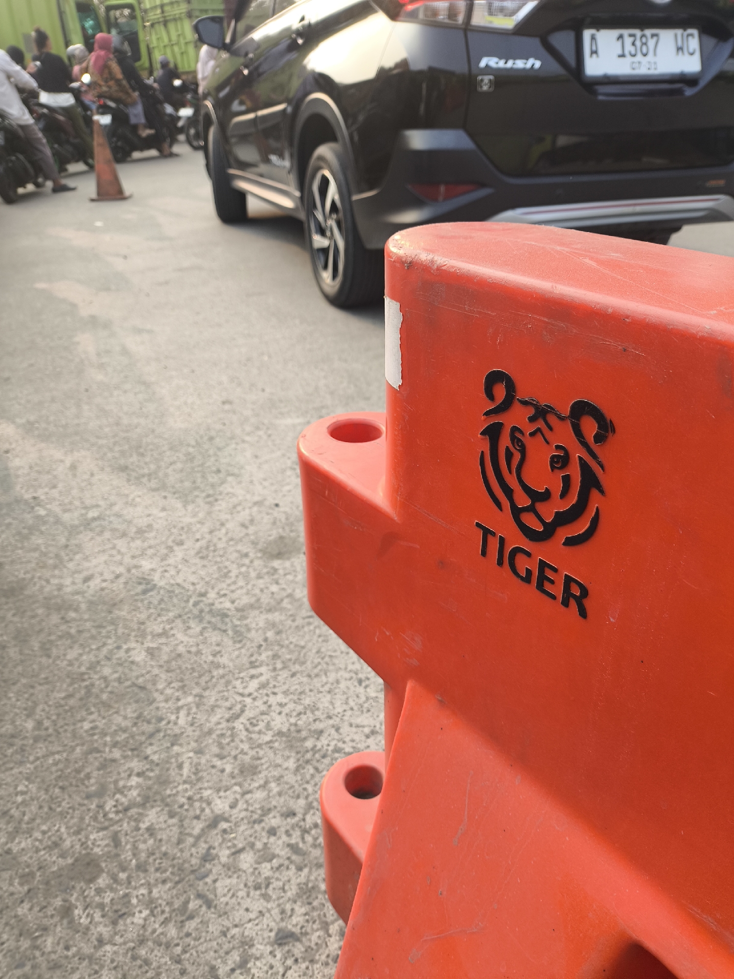 An orange traffic barrier with a black tiger logo and the word "TIGER" printed on it is in the foreground, positioned on a paved surface. In the background, a black car with the license plate "A 1387 WC" is parked. Several people on motorcycles are also visible in the background, some of whom are wearing headscarves. A green truck is partially visible to the left. The scene appears to be on a street in Waringinkurung, Indonesia, likely during daytime due to the lighting conditions.