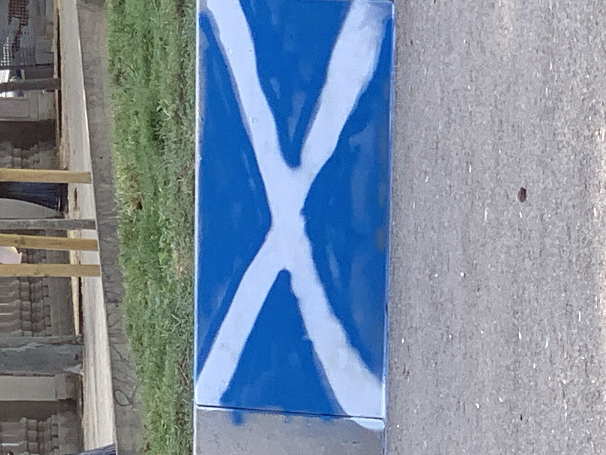 The image displays a close-up of a vertical, rectangular object painted with a blue background and a white Saltire (Saint Andrew's Cross). The paint appears to be spray-painted, with some areas of uneven coverage and drips visible, particularly on the white lines. The object itself seems to be made of a metallic or composite material, with a visible seam or division across the lower portion.
To the left of the painted object, a strip of green grass is visible, suggesting an outdoor setting. Beyond the grass, there are indications of a building or structure with horizontal wooden beams and what appear to be stacked bricks or concrete blocks. These elements are partially obscured and out of focus. The background on the right side of the painted object is a textured gray surface, likely asphalt or concrete, with small white flecks and a few dark specks.
The lighting suggests it is daytime, possibly late afternoon or early morning, given the quality of the light and shadows, though no distinct shadows are prominently cast. The weather appears to be fair. The image is a still shot, with no discernible activity taking place beyond the static presentation of the painted object. There is no visible text in the image. The context provided is Barcelona, Spain, which is not directly evident from the visual elements alone, but might inform interpretations of the object's purpose or significance if it were a known landmark or symbol within that location.